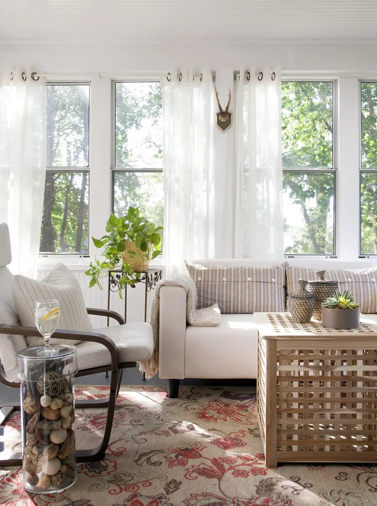 Sunlit sitting room designed by Georgia Zikas featuring sheer white curtains, a striped sofa, floral area rug, woven coffee table, and neutral accent chair layered with natural textures and botanical details.