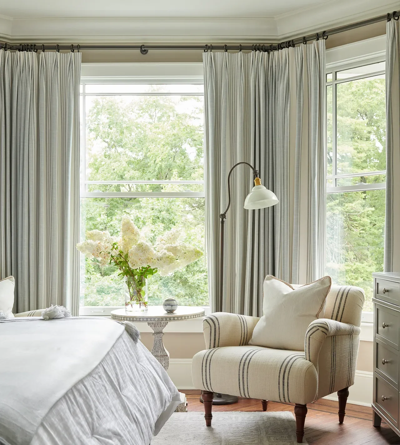 Light-filled bedroom corner with soft neutral curtains, a striped armchair, side table with flowers, and large windows creating a calm and inviting seating area.