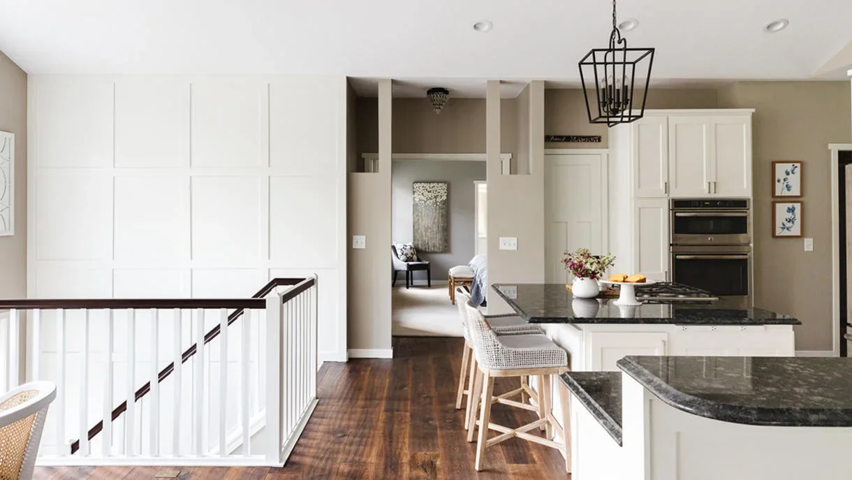Open-concept kitchen and dining area with white paneled accent wall, dark wood floors, granite island with woven barstools, and black lantern pendant lighting.