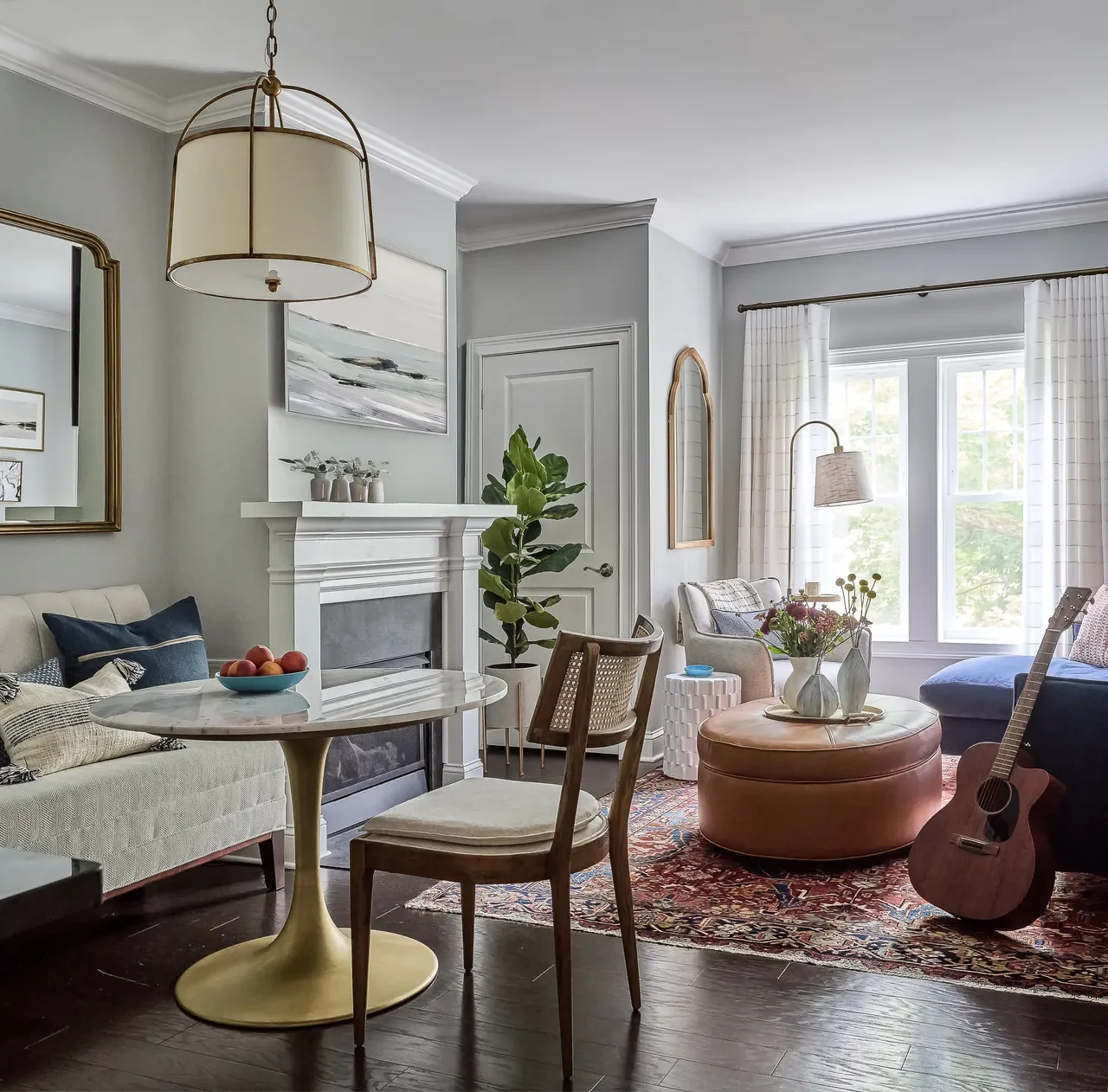 Small living room designed by Georgia Zikas featuring a white fireplace, round leather ottoman coffee table, marble dining table, patterned rug, and large windows that bring in natural light.