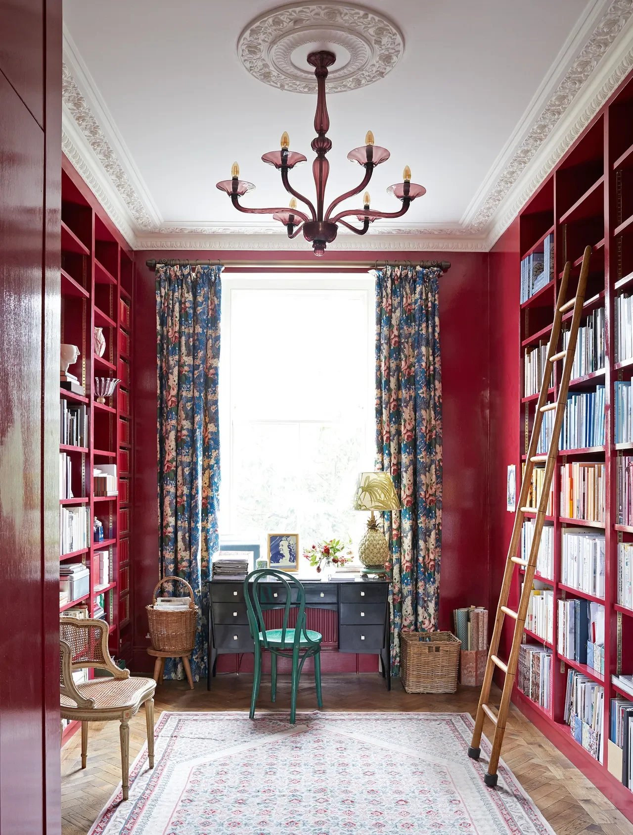 Custom home office and library designed by Georgia Zikas featuring rich red built-in bookshelves, a rolling library ladder, floral drapery, decorative crown molding, and a desk centered beneath a large window.