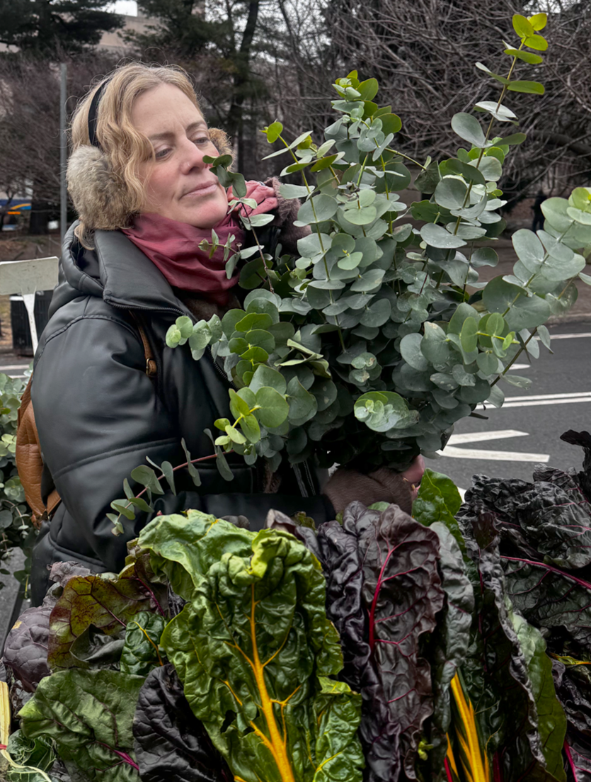 A photo of Jessie White, founder of Harness Whole Health Functional Nutrition, holding a eucalyptus branches at a farmers market, with fresh leafy green vegetables in the foreground.