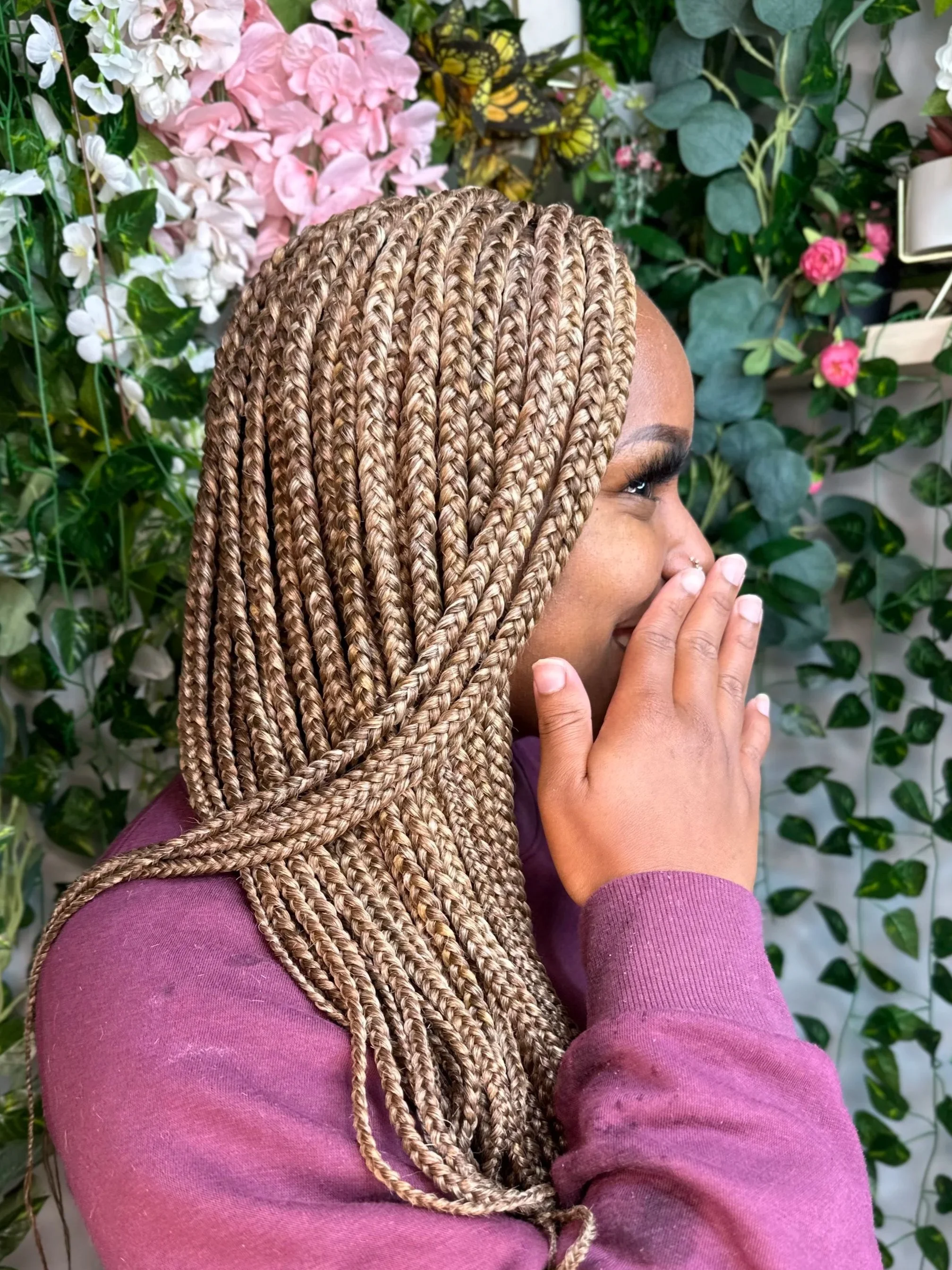 A woman with blonde box braids wearing a purple long-sleeve shirt, standing in front of a wall of green leaves and pink and white flowers, smiling with her eyes closed and hand near her face.