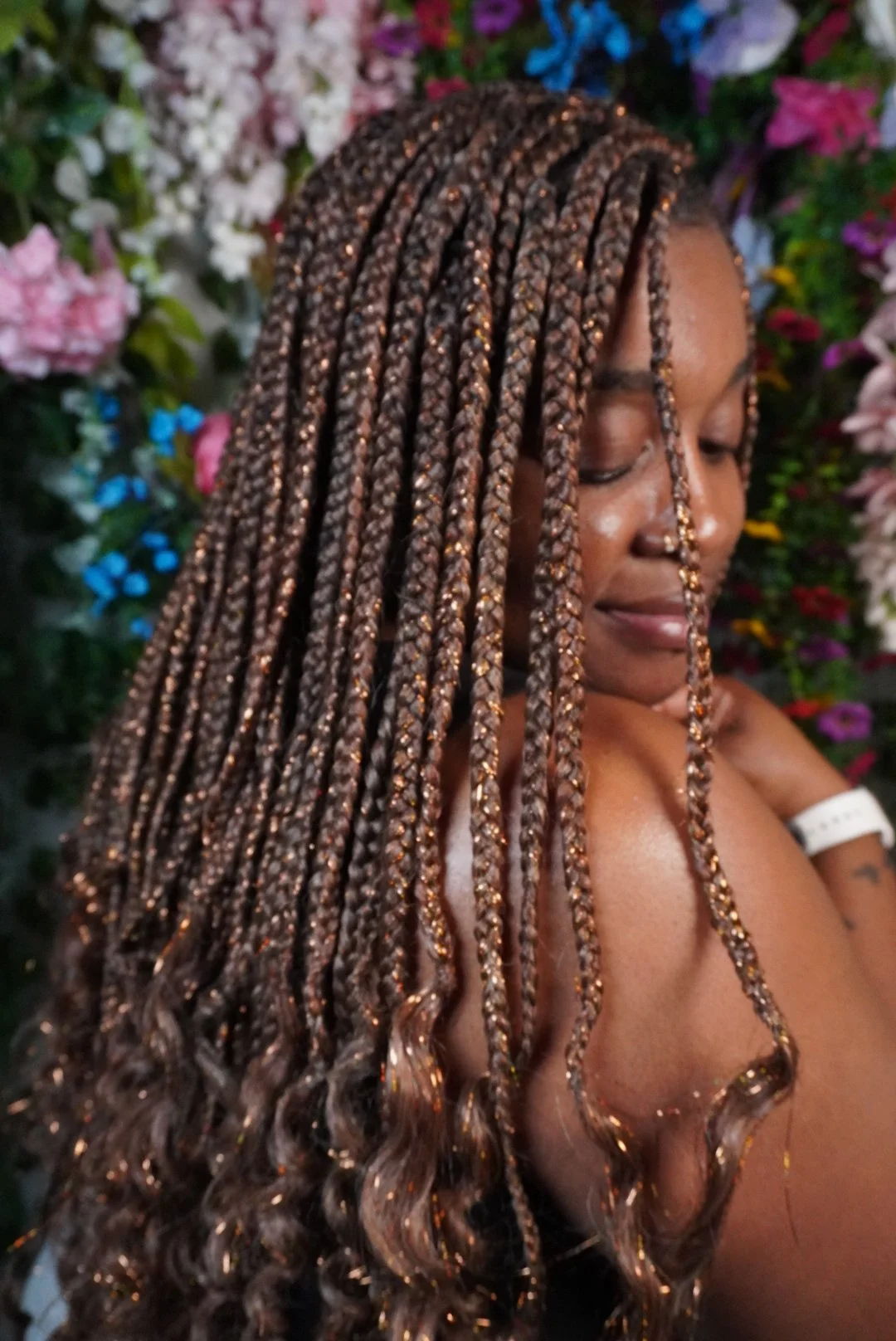A woman with braided hair with golden accents, standing against a background of colorful flowers, smiling with her eyes closed.