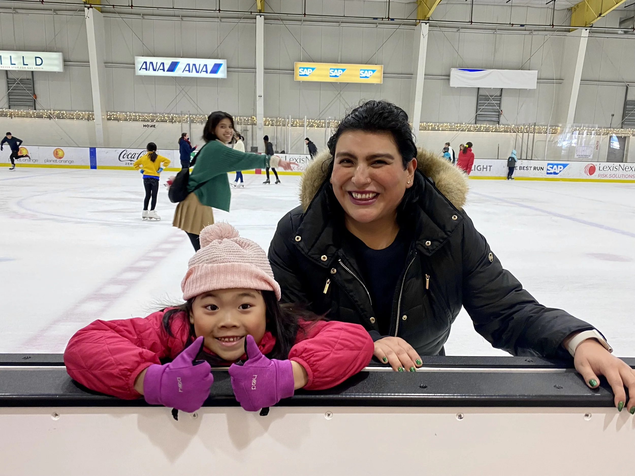 A Figure Skating Coach and her young student in pink coats and purple gloves sitting at an ice skating rink, smiling at the camera, with other skaters in the background.