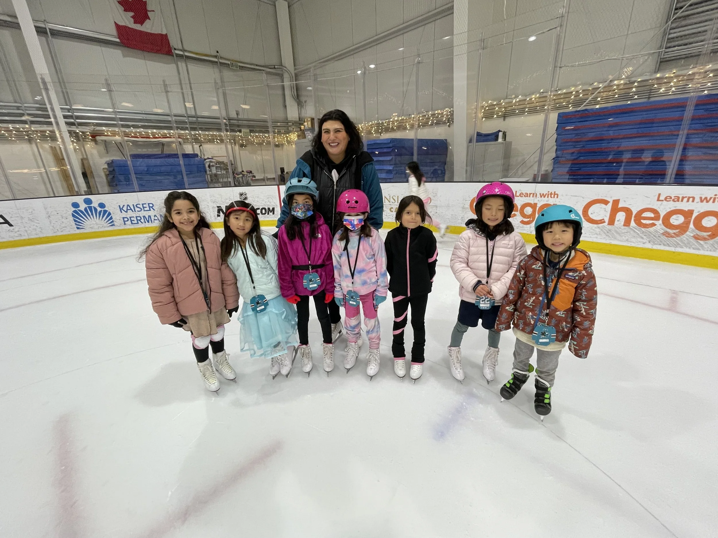 A group of children in colorful winter clothing and helmets, standing on ice skates at an indoor ice skating rink, with a smiling Figure Skating Coach among them in San Jose, CA.