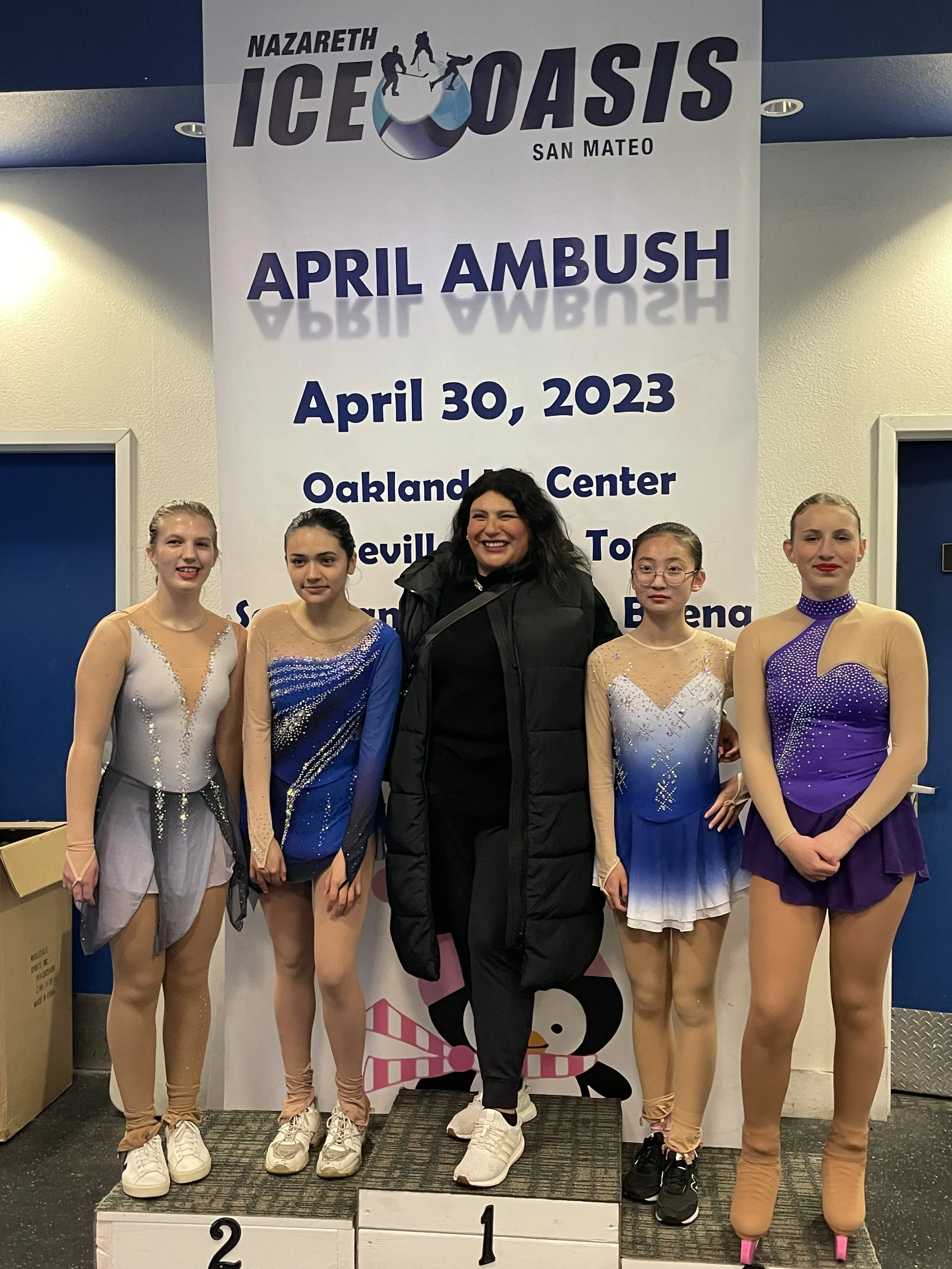 A group of five female figure skaters and their coach stand on a winners' podium, smiling after a competition. The backdrop displays a banner for the Nazareth Ice Oasis in San Mateo with the event name 'April Ambush' dated April 30, 2023.