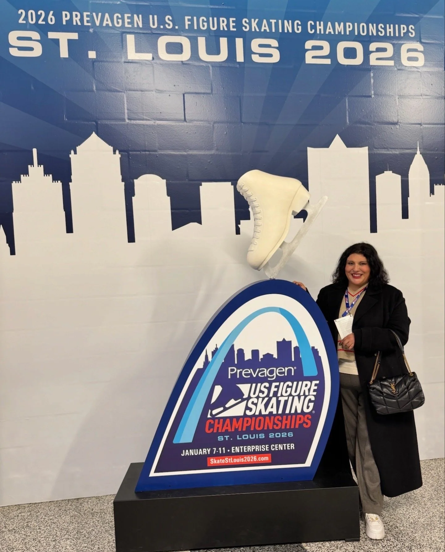 Figure Skating Coach standing next to a sign for the 2026 Prevalent US Figure Skating Championships in St. Louis, January 7-11, 2026, at the Enterprise Center. The sign features the St. Louis city skyline silhouette.