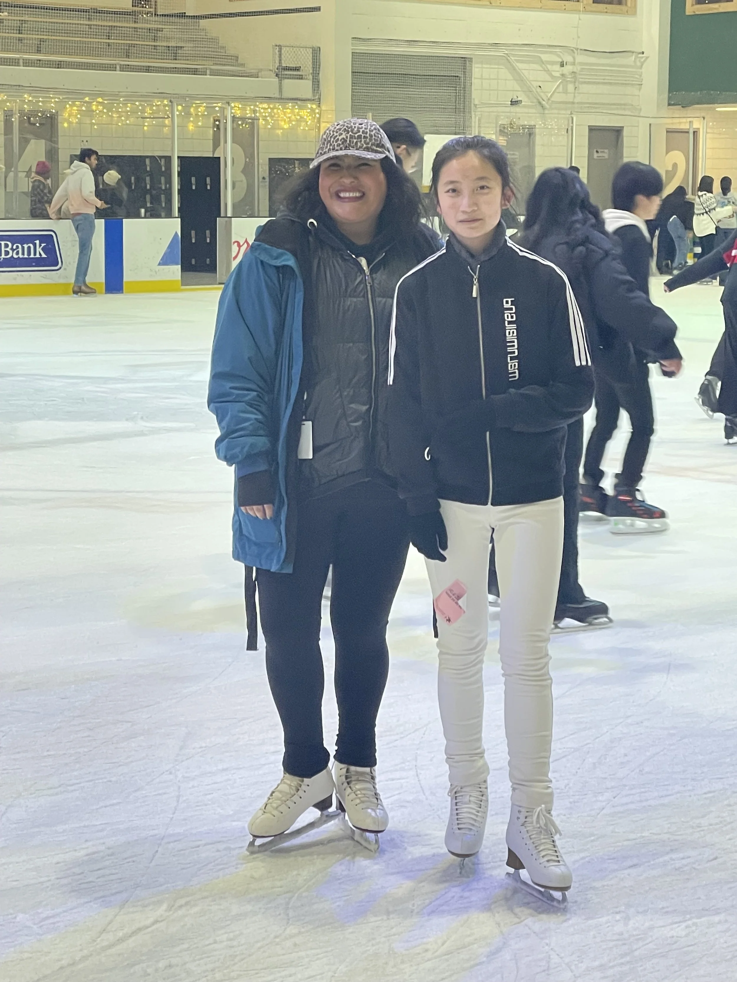 Two women ice skating at an indoor rink, smiling at the camera. One wearing a leopard print hat and black jacket, the other in a black and white zip-up jacket and white pants. Several other skaters are visible in the background.