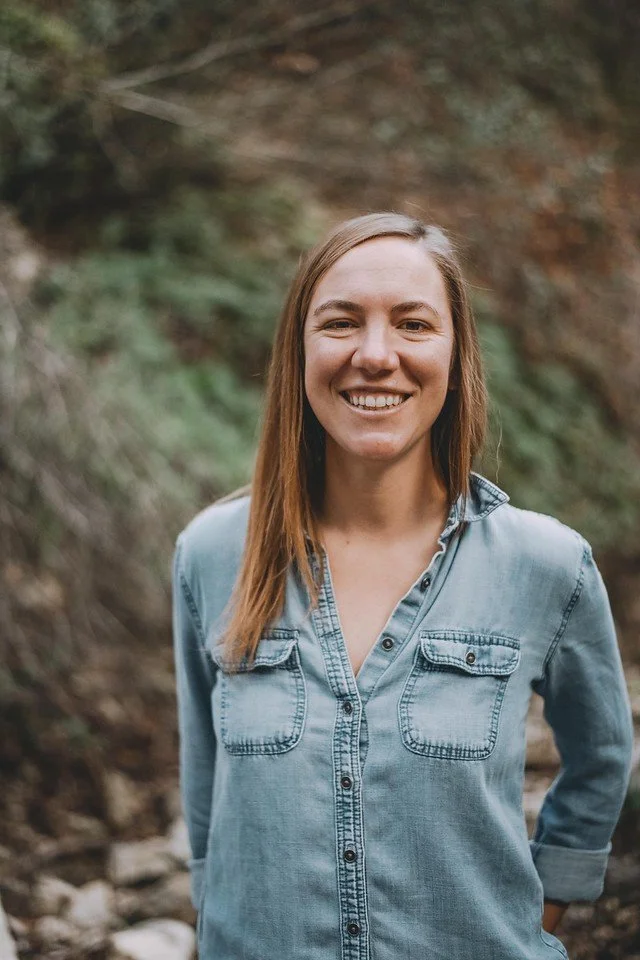 Smiling woman in denim shirt standing outdoors in a wooded area.