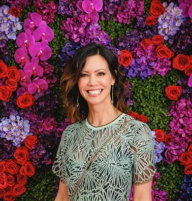 A woman smiling in front of a colorful floral backdrop with pink, red, purple, and green flowers.