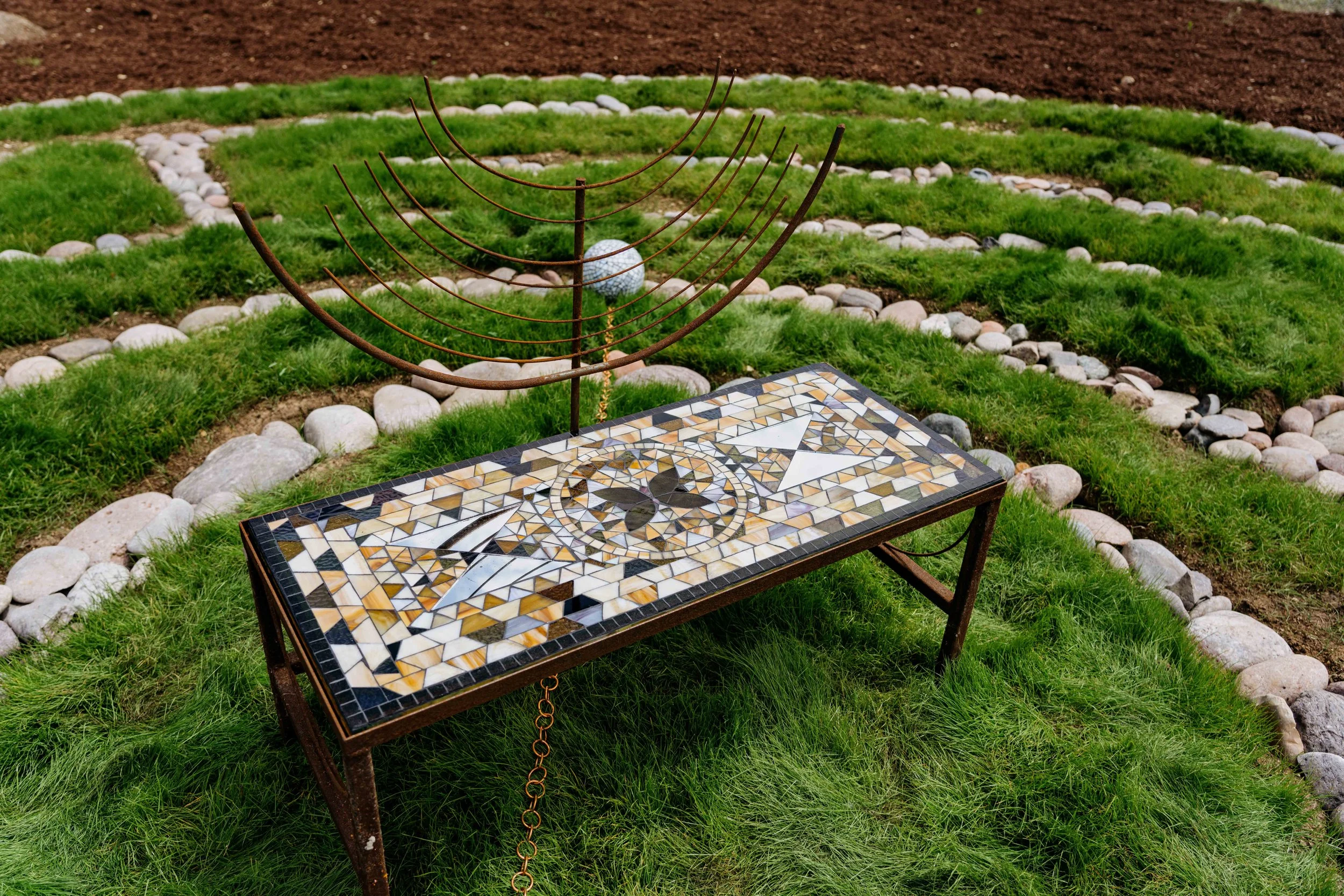 A mosaic table with sundial and a metal sculpture of a globe on a landscaped lawn with grass, rocks, and flower beds.