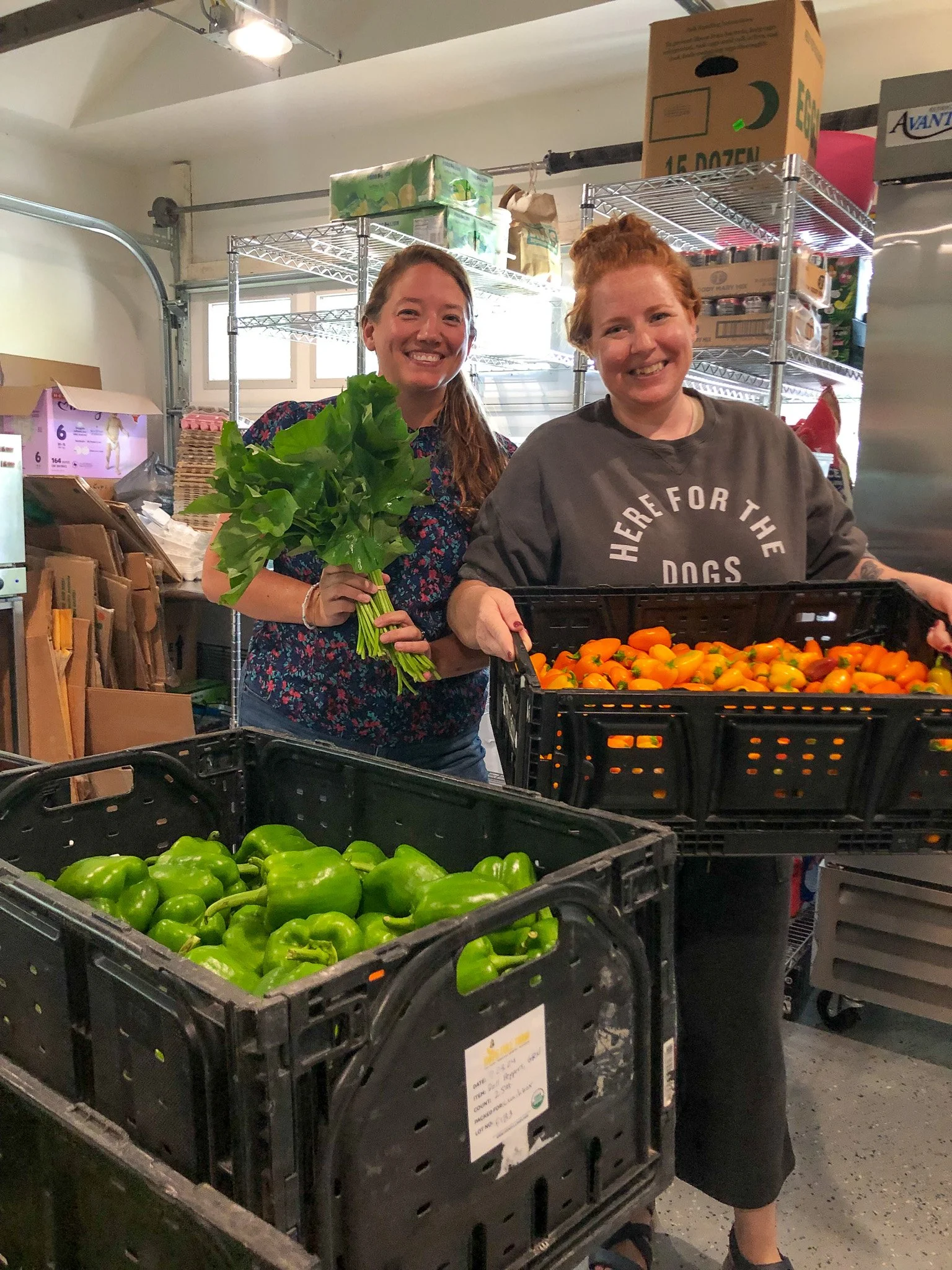 Two women smiling in at sunday lunchbox, one holding a bunch of green leafy vegetables, with crates of green bell peppers and orange peppers in front of them.