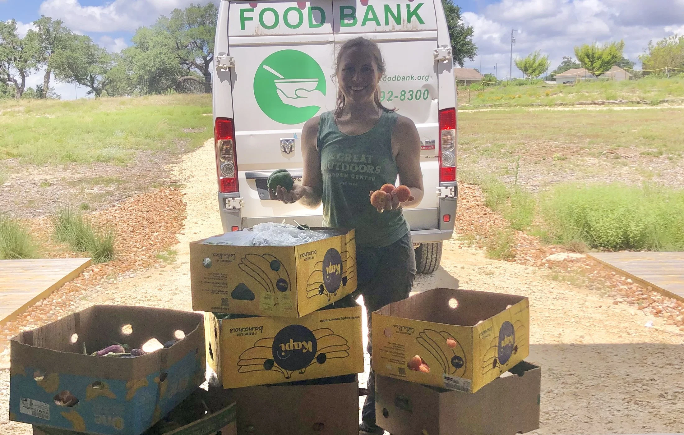 Woman standing in front of a food bank vehicle holding produce, with four boxes of fruit and vegetables in front of her.