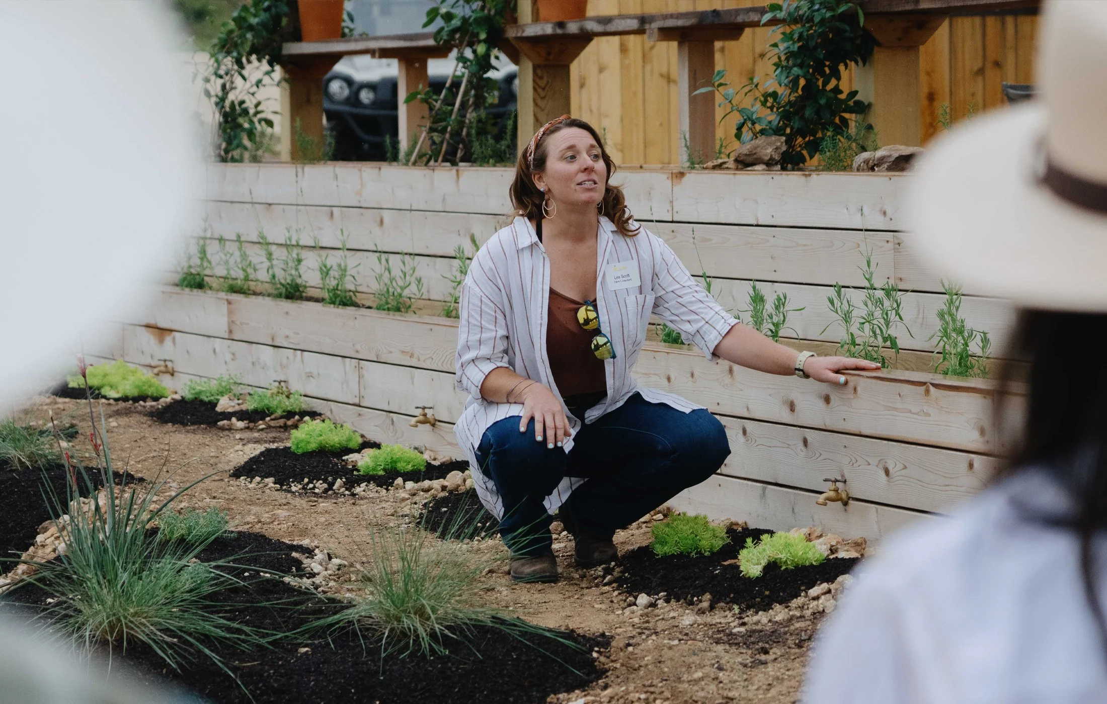 A woman crouching next to a wooden garden bed, speaking to a group, with plants and gardening taps visible in the background.