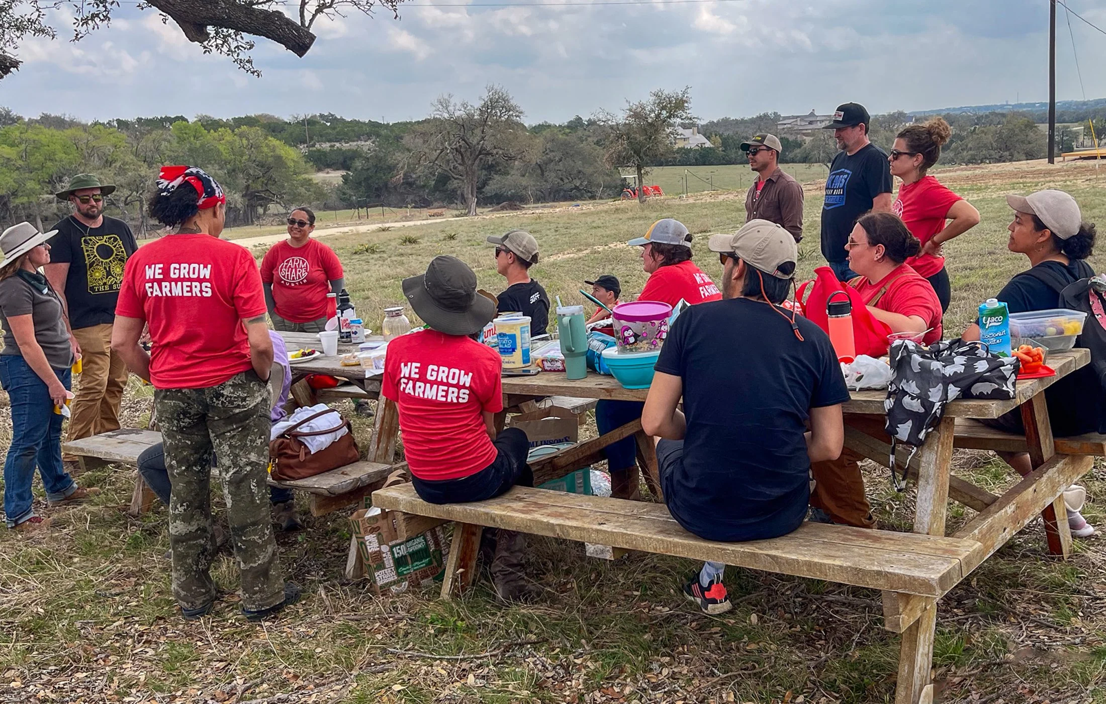 A group of people gathered outdoors around a wooden picnic table, some wearing red shirts with the words "We Grow Farmers," enjoying a social event in a rural setting with trees and open fields in the background.