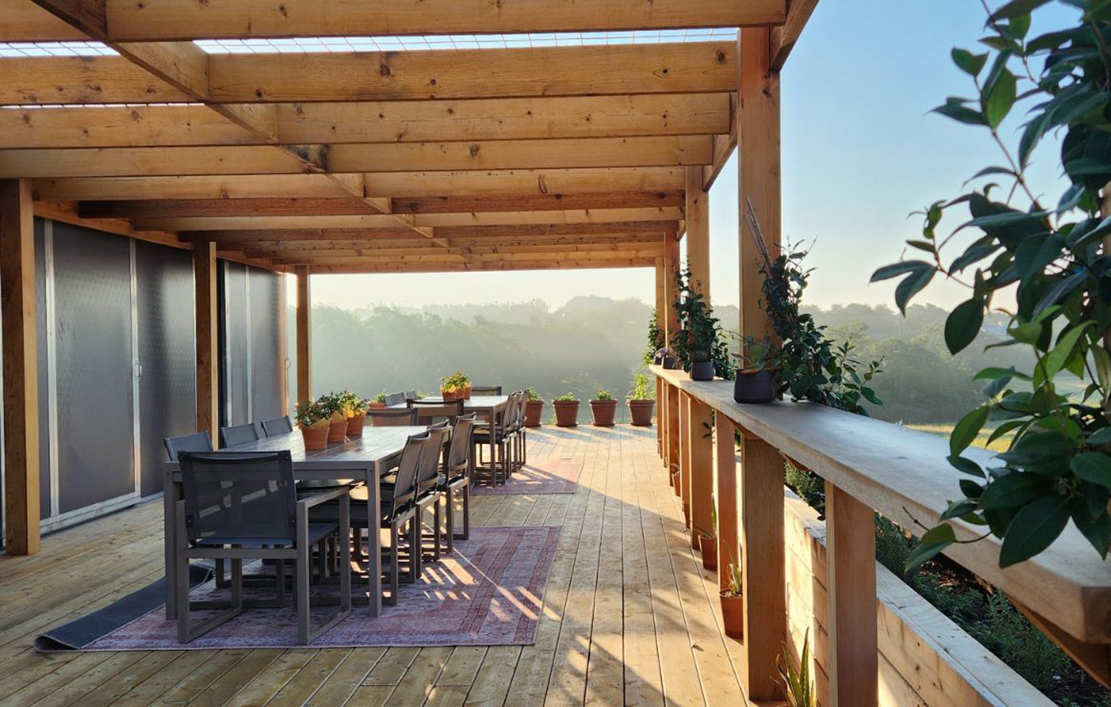 Outdoor wooden deck with dining table, chairs, potted plants, and a scenic view of trees in the distance.