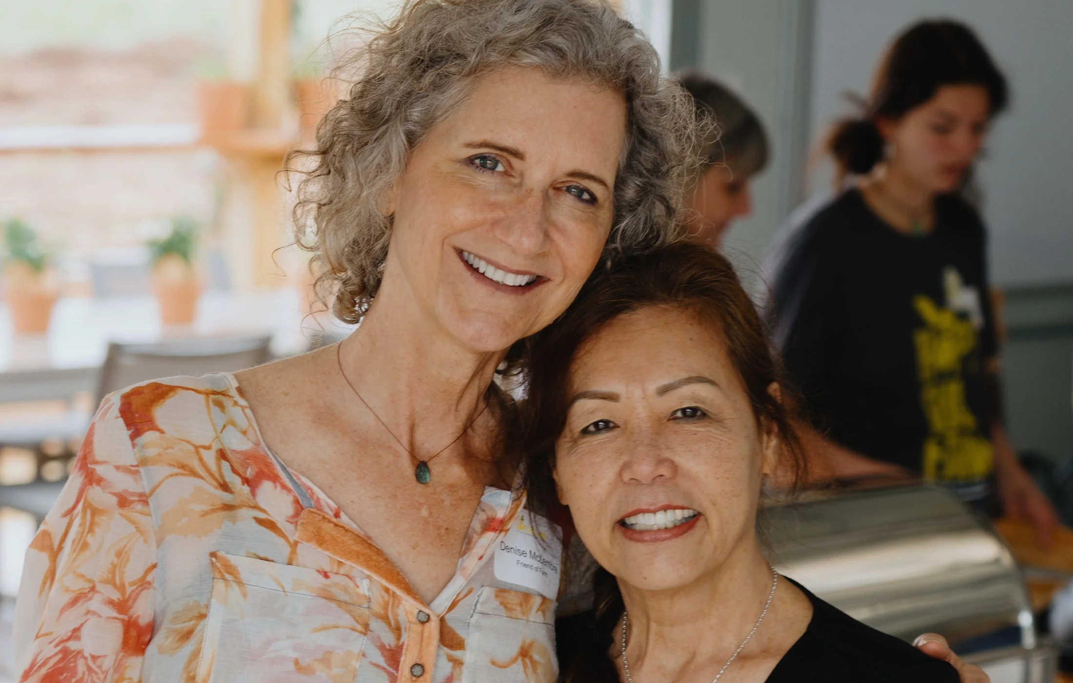 Two women smiling and posing for a photo indoors, with other people in the background.