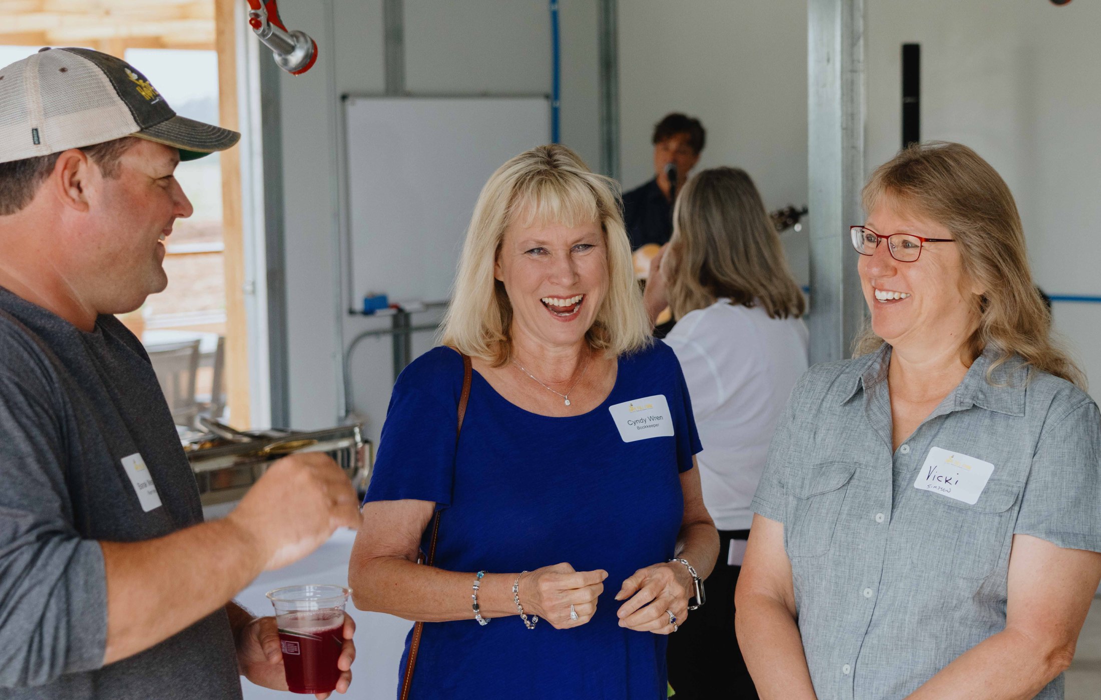 Three people smiling and engaging in conversation at a social gathering, with a man on the left holding a drink and two women in the center and right wearing name tags, inside a bright room with a band playing in the background.