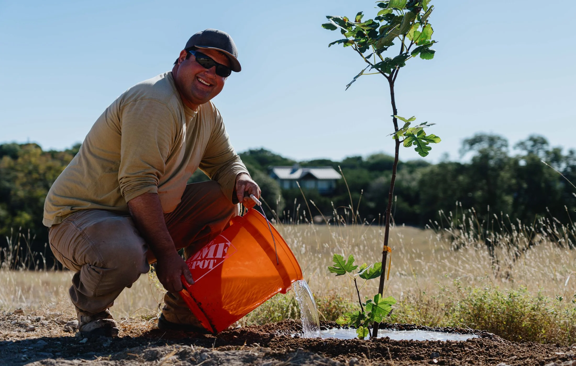 A man planting a small tree, pouring water from an orange Home Depot bucket in a sunny outdoor field.
