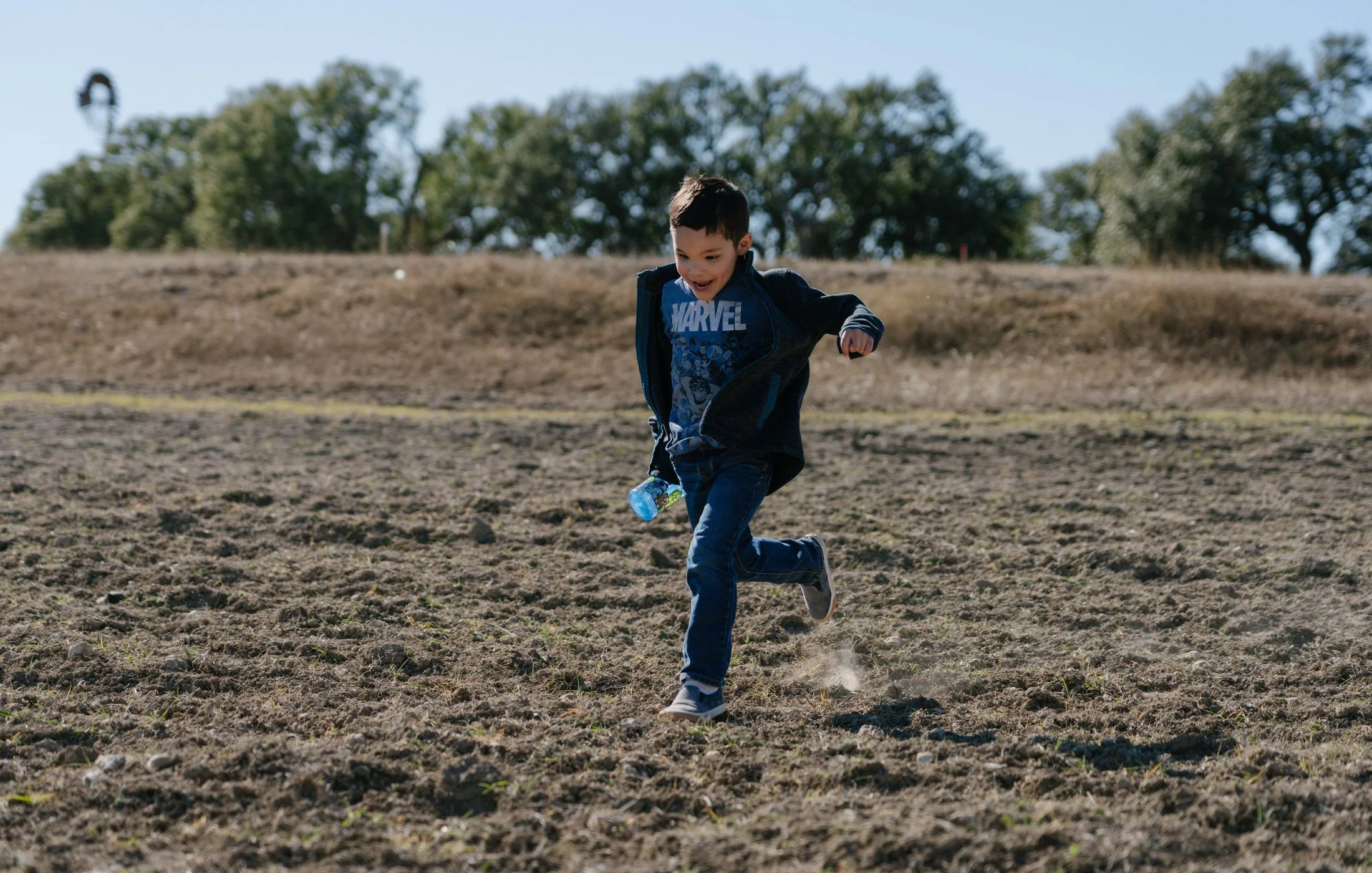A young boy in a Marvel shirt running across a dry, dirt field with trees in the distance.