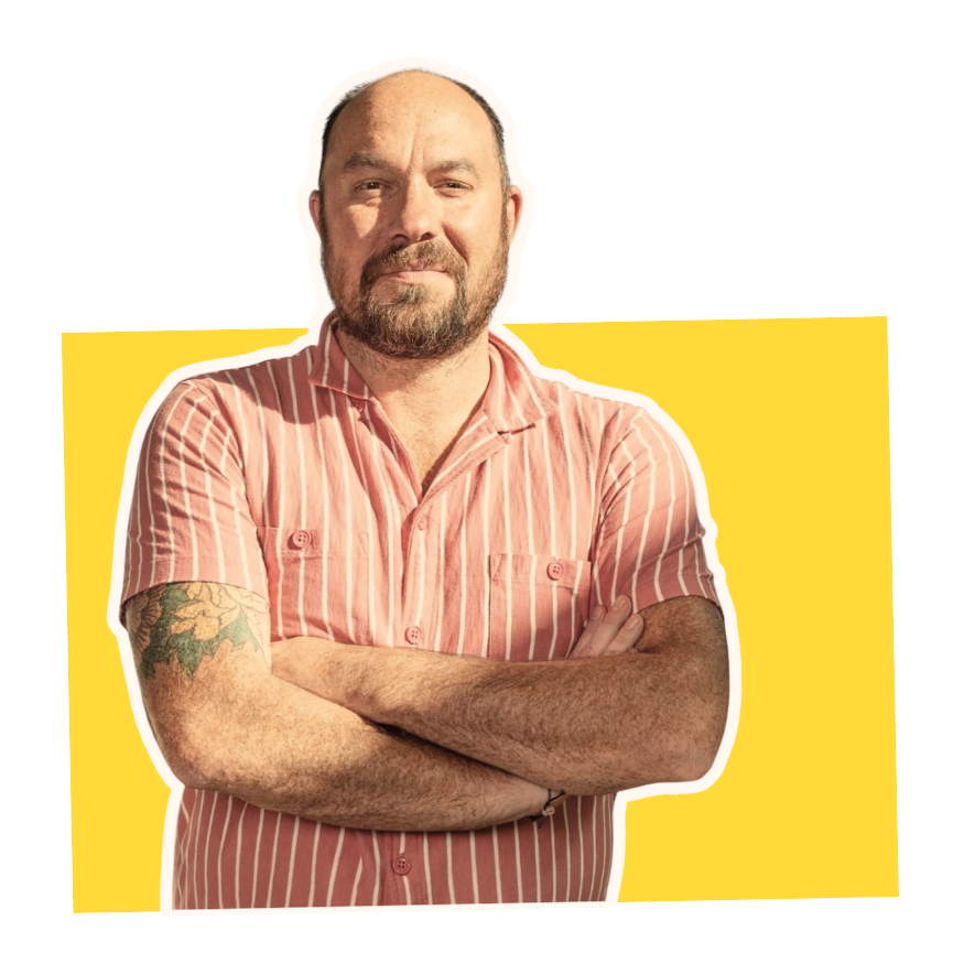 A man with a beard and tattooed arm, wearing a pink and white striped shirt, standing with arms crossed against a colorful background of yellow, red, and black sections.