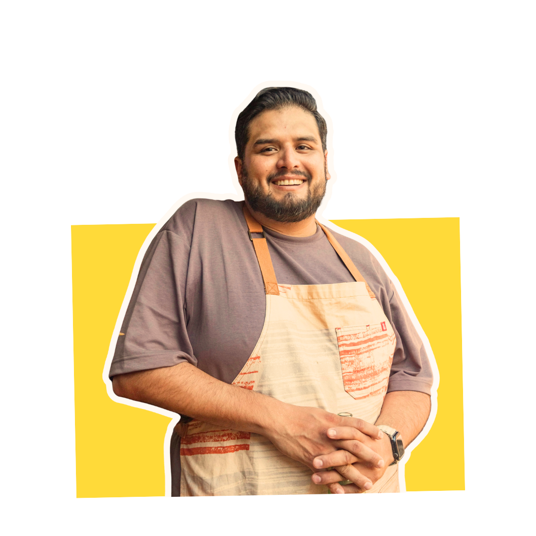 Smiling man with a beard wearing a gray t-shirt and an apron, standing against a yellow and white background.