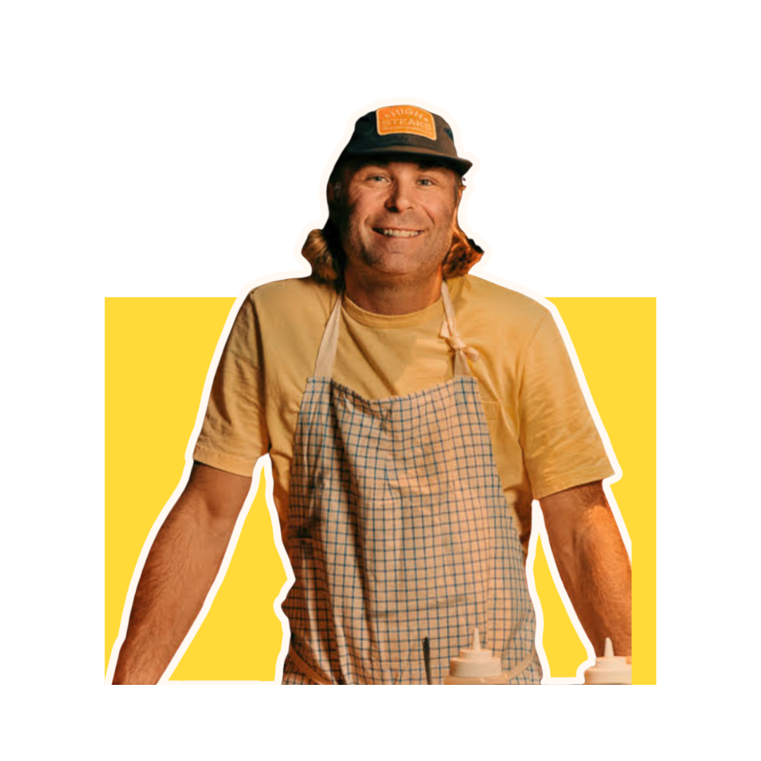 A man wearing a cap, apron, and beige T-shirt, smiling, standing behind a counter with condiments.