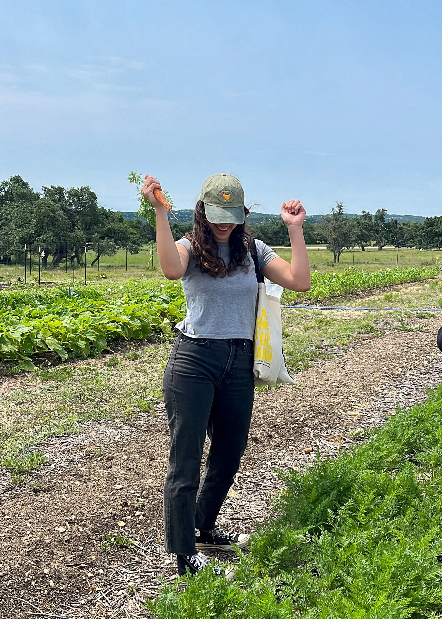 A young woman standing in a farm field holding a carrot in her right hand, wearing a gray t-shirt, black jeans, a beige cap, and black sneakers, smiling and celebrating.
