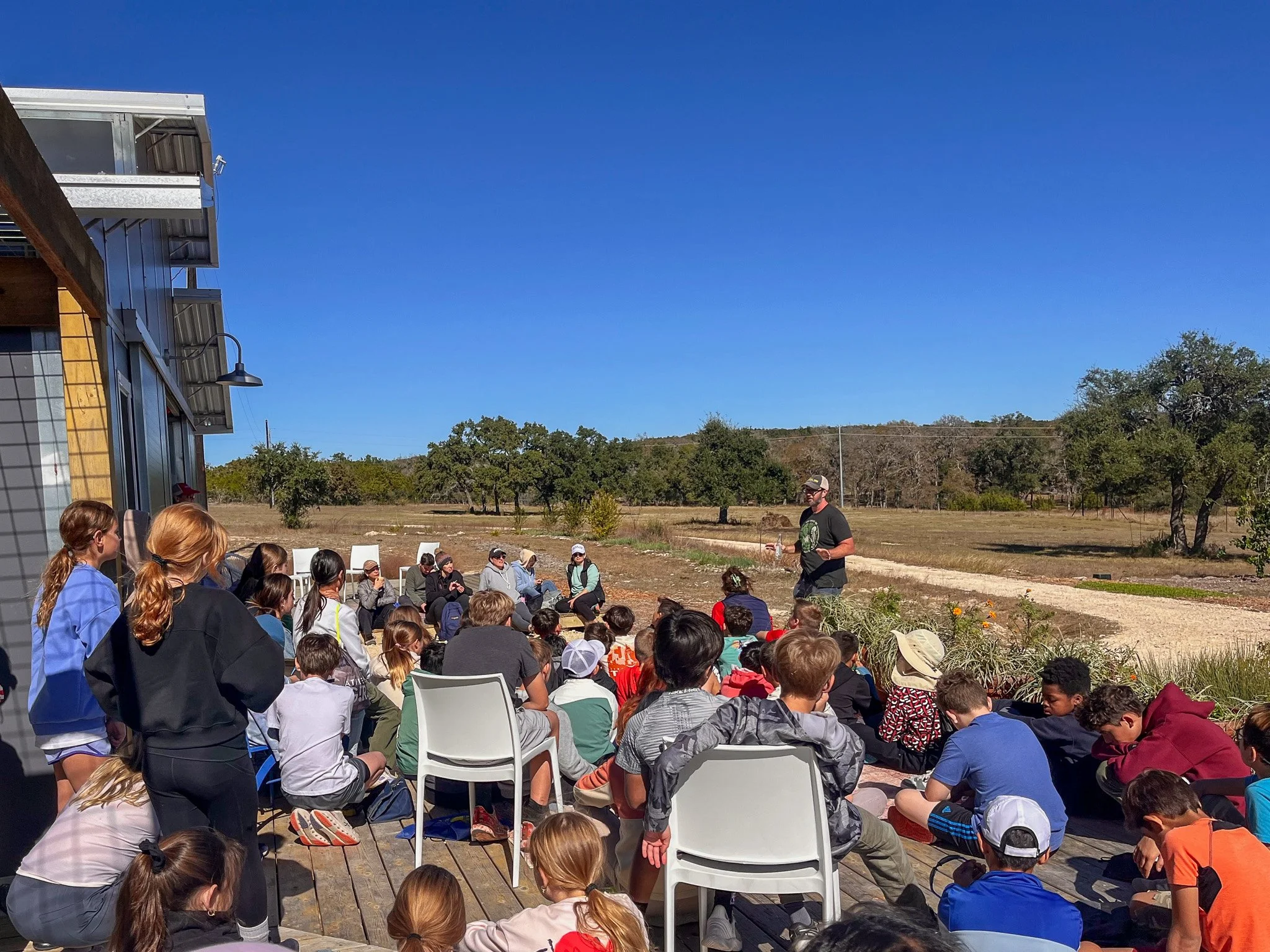 Group of children and adults sitting outdoors on a wooden deck, listening to a man standing in front of a rural landscape with trees and a dirt road, during a sunny day.