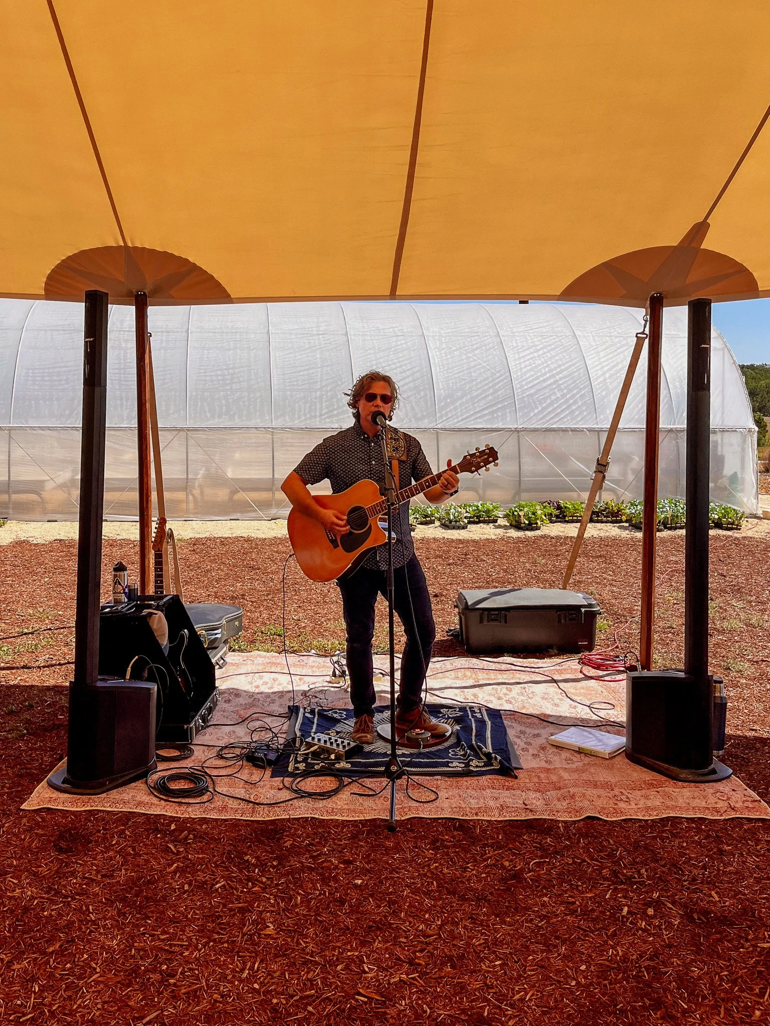 A person playing guitar and singing into a microphone on an outdoor stage with a tent overhead and greenhouse in the background.