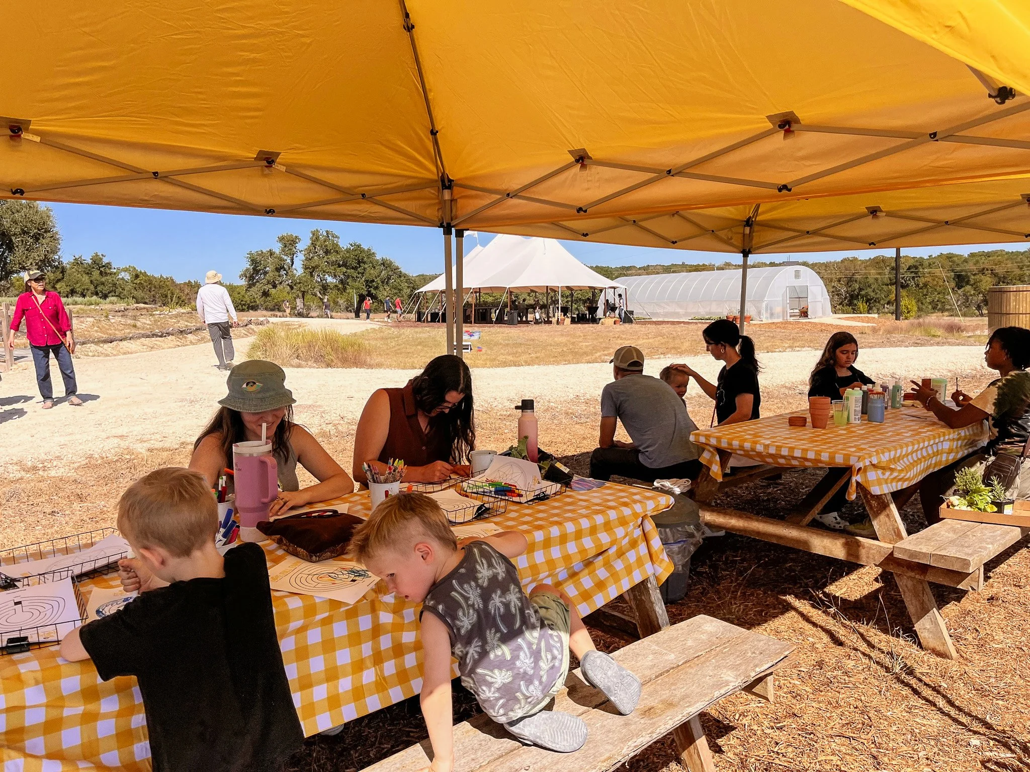 People sitting at tables under a yellow canopy outdoors, with some children and adults, engaging in arts and crafts activities, with a white tent and greenhouses in the background.
