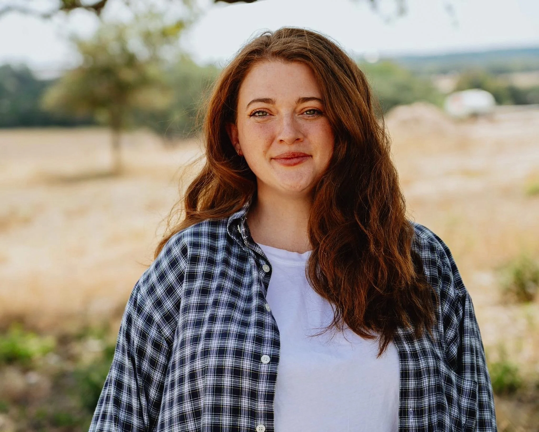 A young woman with wavy brown hair wearing a blue plaid shirt with trees and a grassy landscape in the background.