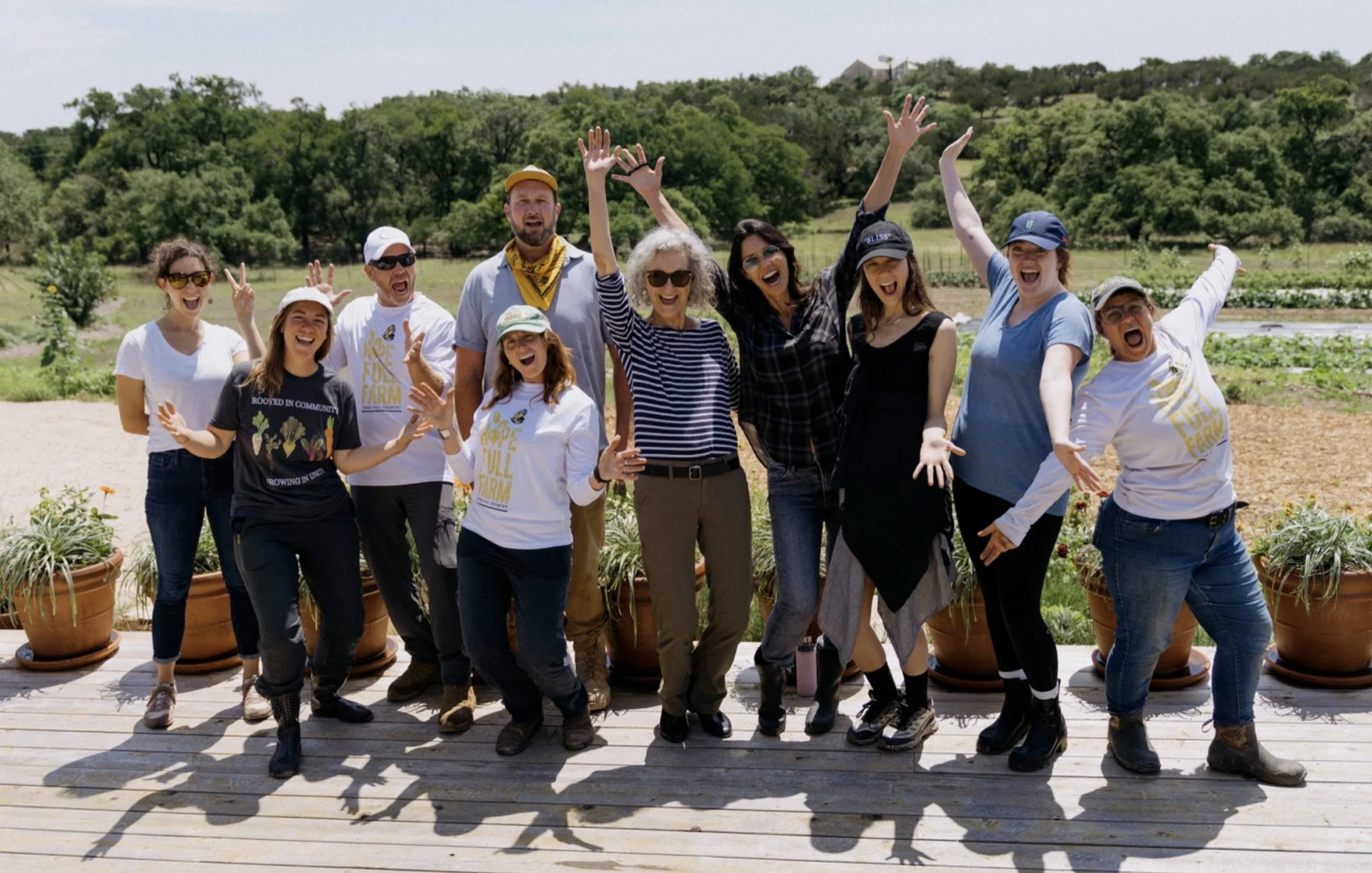 A group of people standing outdoors on a deck, smiling and cheering, with a scenic green landscape and hills in the background.