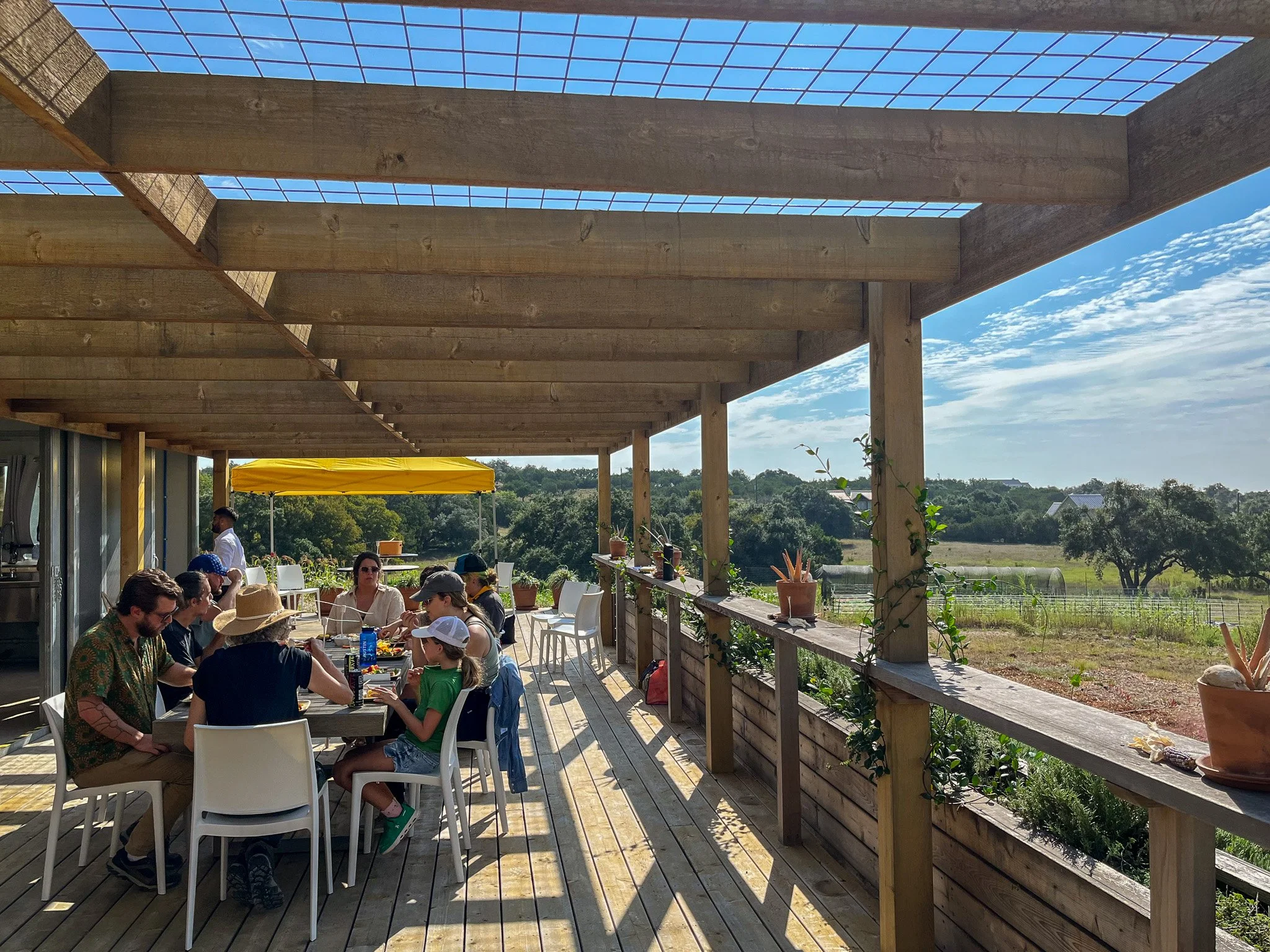 People sitting at a long table on a wooden outdoor patio with a view of trees and blue sky, some wearing hats, during daytime.