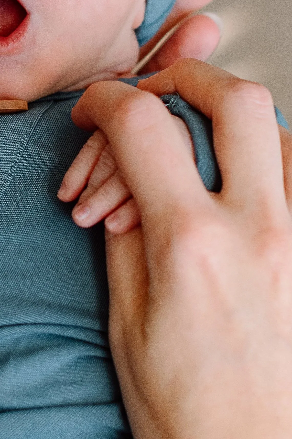 A newborn baby boy in matching pale blue onesie and hat yawns while being cradled by parents hands