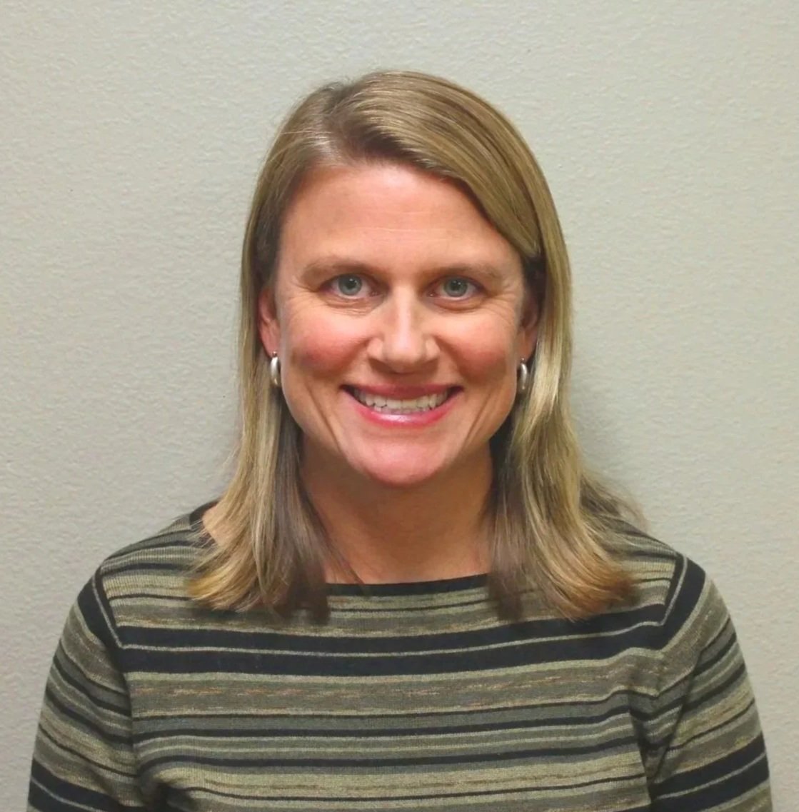 Portrait of a woman with long red hair wearing a striped shirt smiling against a gray background.