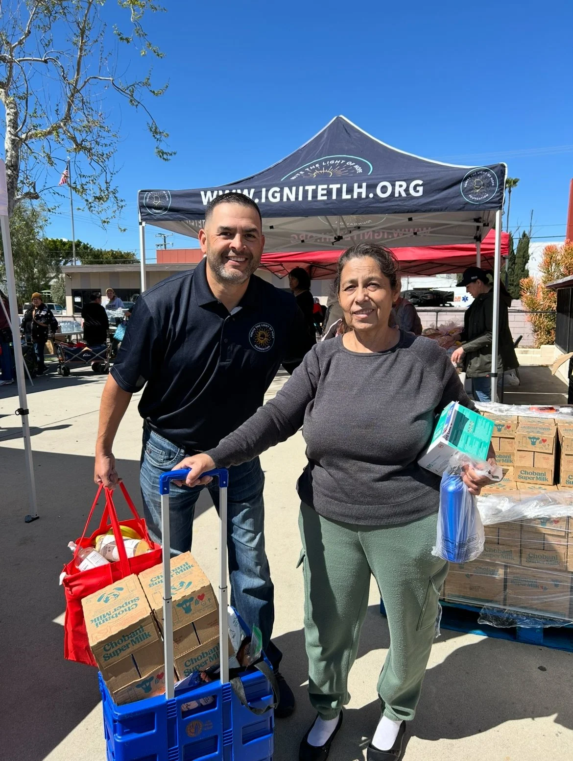 A man and a woman at a community food distribution event, standing in front of a black tent with the website 'IGNITE LTH.org' printed on it. The woman is holding groceries and the man is pushing a blue cart filled with boxes and bags of food. There are other people and tables behind them.