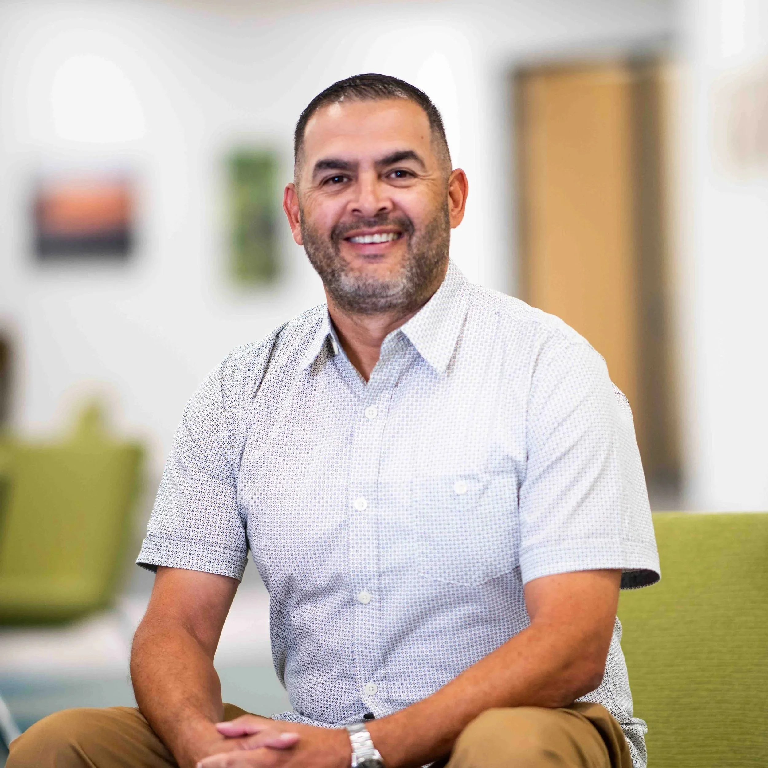 A man with short dark hair and a beard, wearing a dark suit jacket and white shirt, smiling at the camera in front of a purple, patterned background.