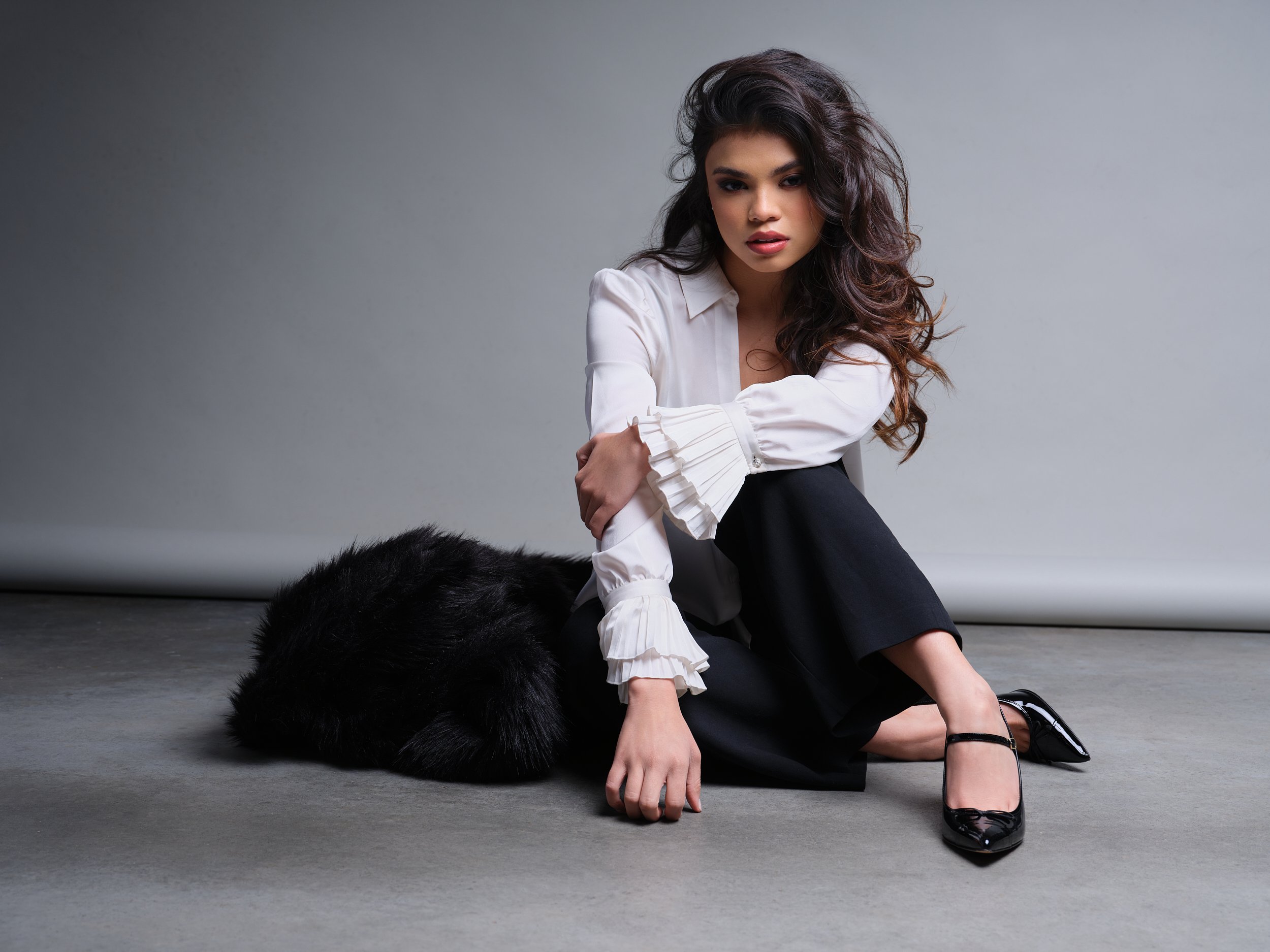 A woman with dark, wavy hair, sitting on the floor against a plain gray background, wearing a white blouse with ruffled details on the sleeves and black pants. She is posed with one knee up and the other leg extended, with her arm resting on her knee and her hand near her face. She is wearing black high heels with ankle straps and looks directly at the camera with a neutral expression.