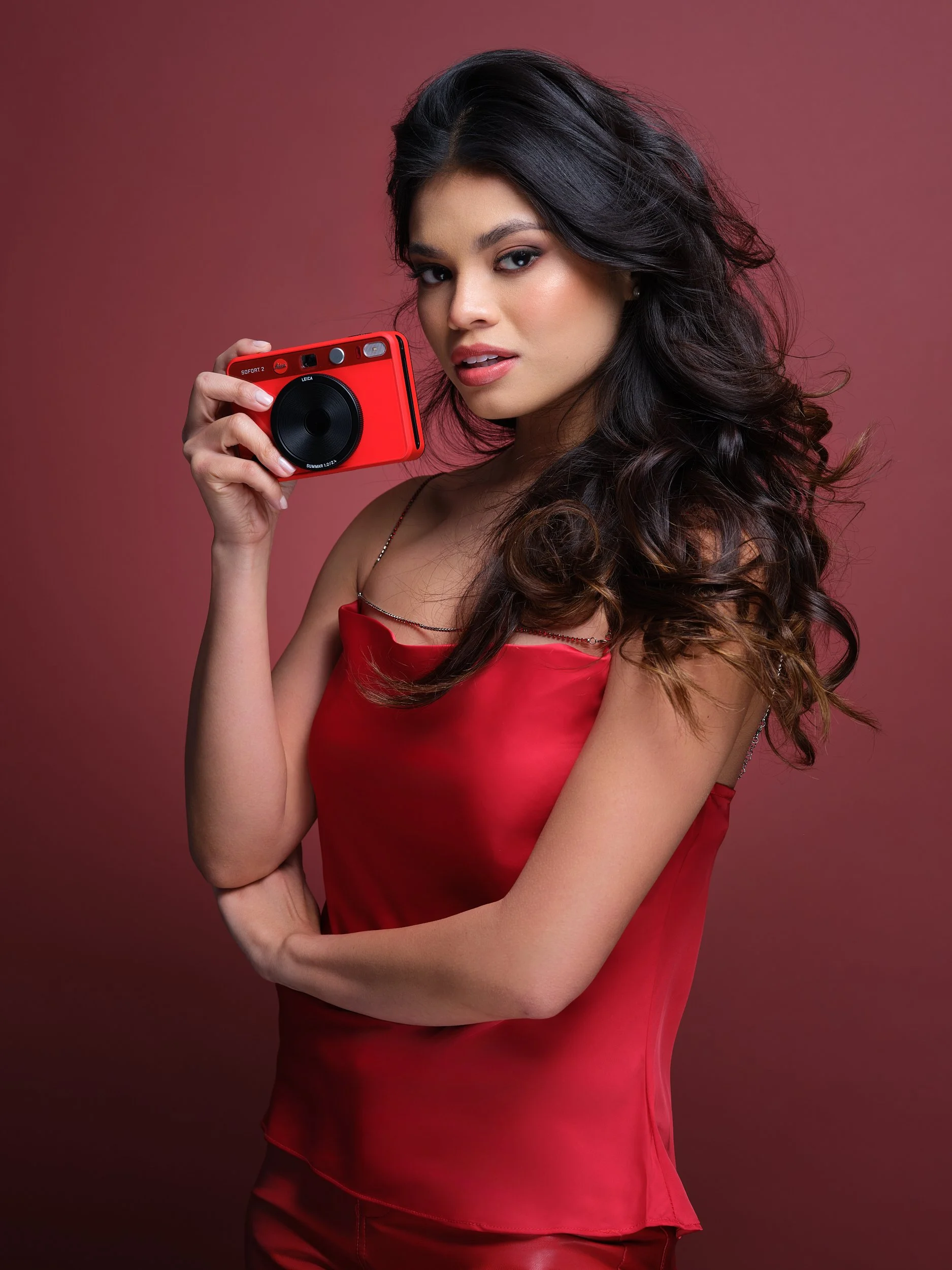 A woman with dark, curly hair and light skin holding a red camera, wearing a red dress, against a pink background.
