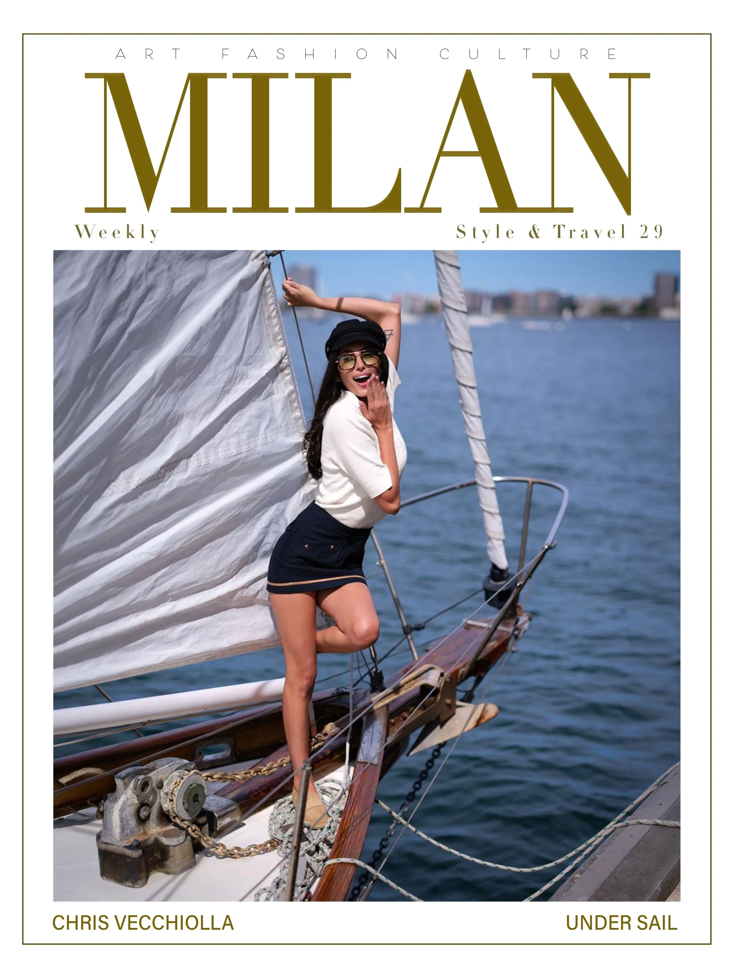 A woman with long hair, sunglasses, a black cap, a white shirt, and dark shorts stands on a sailboat, smiling and touching her face, with her left hand raised holding a rope. The sailboat has white sails, and the background shows water and blurred ci