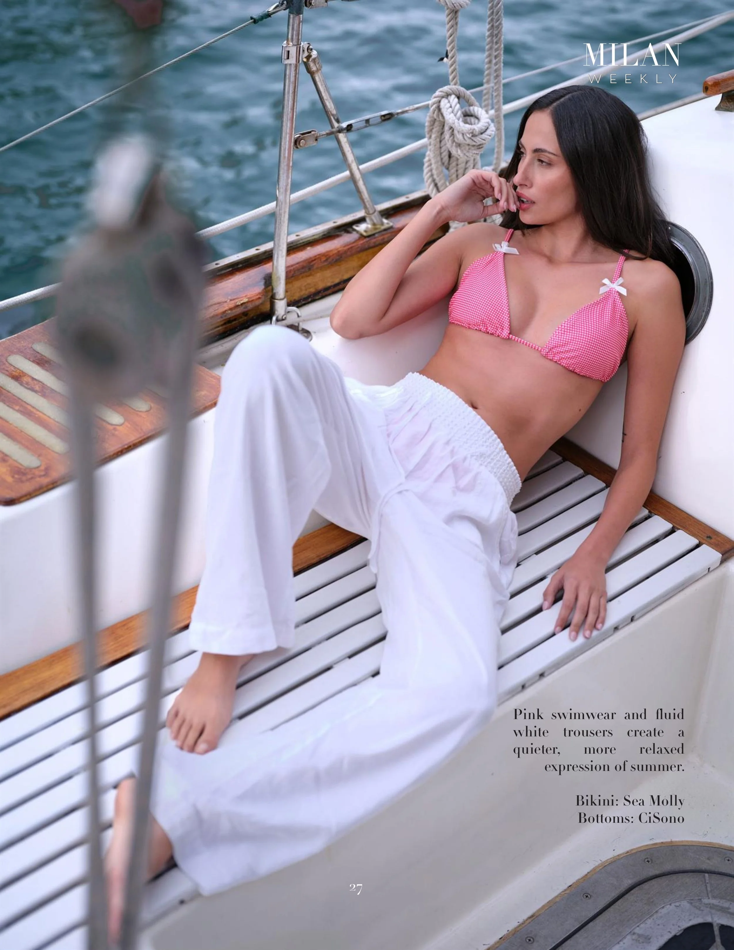 A woman in pink swimsuit and white trousers relaxing on a boat, with its deck made of white slats and wooden accents, surrounded by water with visible boat rigging.