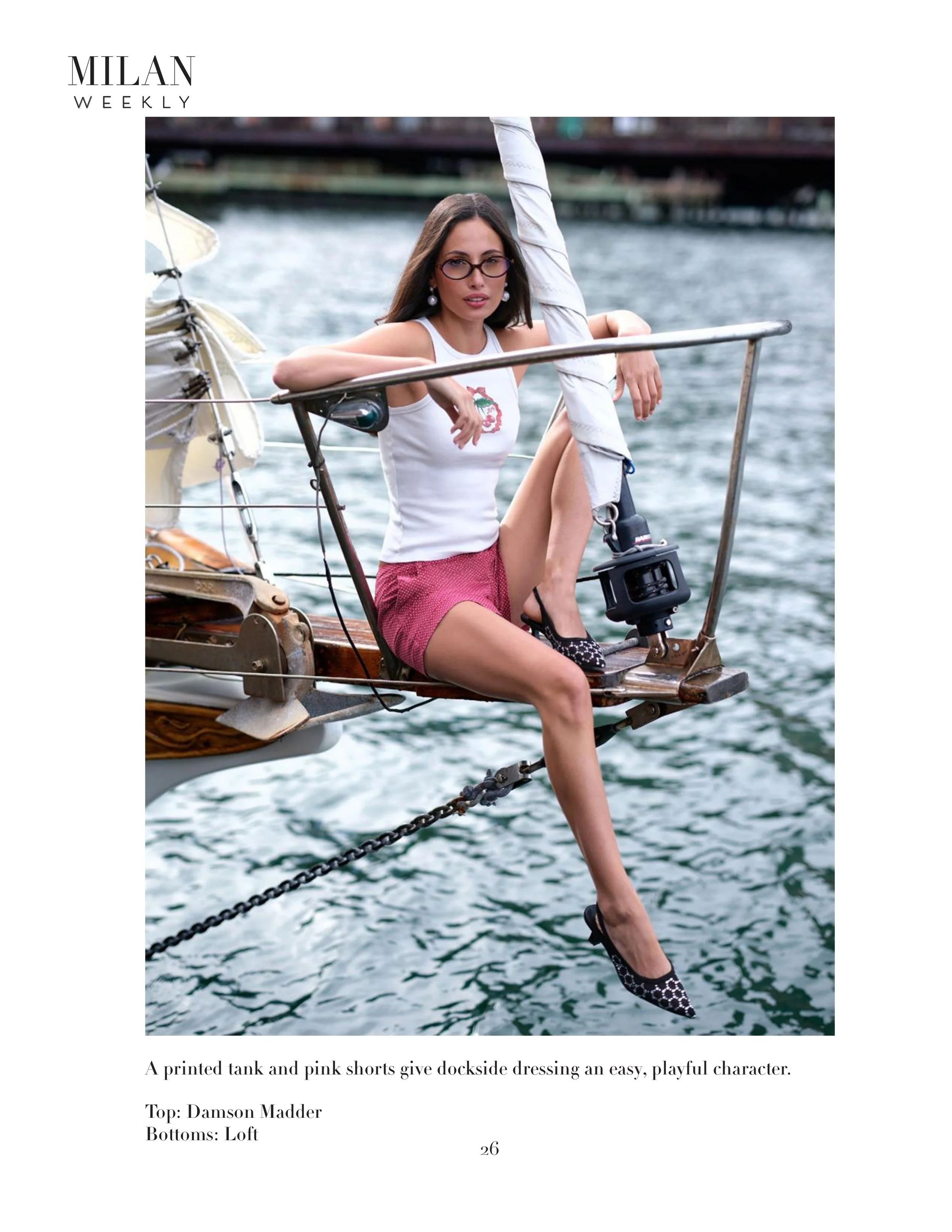 A woman dressed in a white tank top and pink shorts, sitting on the bow of a boat. She wears glasses and earrings, with her left arm resting on a metal railing and her right arm raised above her head. The boat is in the water, with a cityscape in the