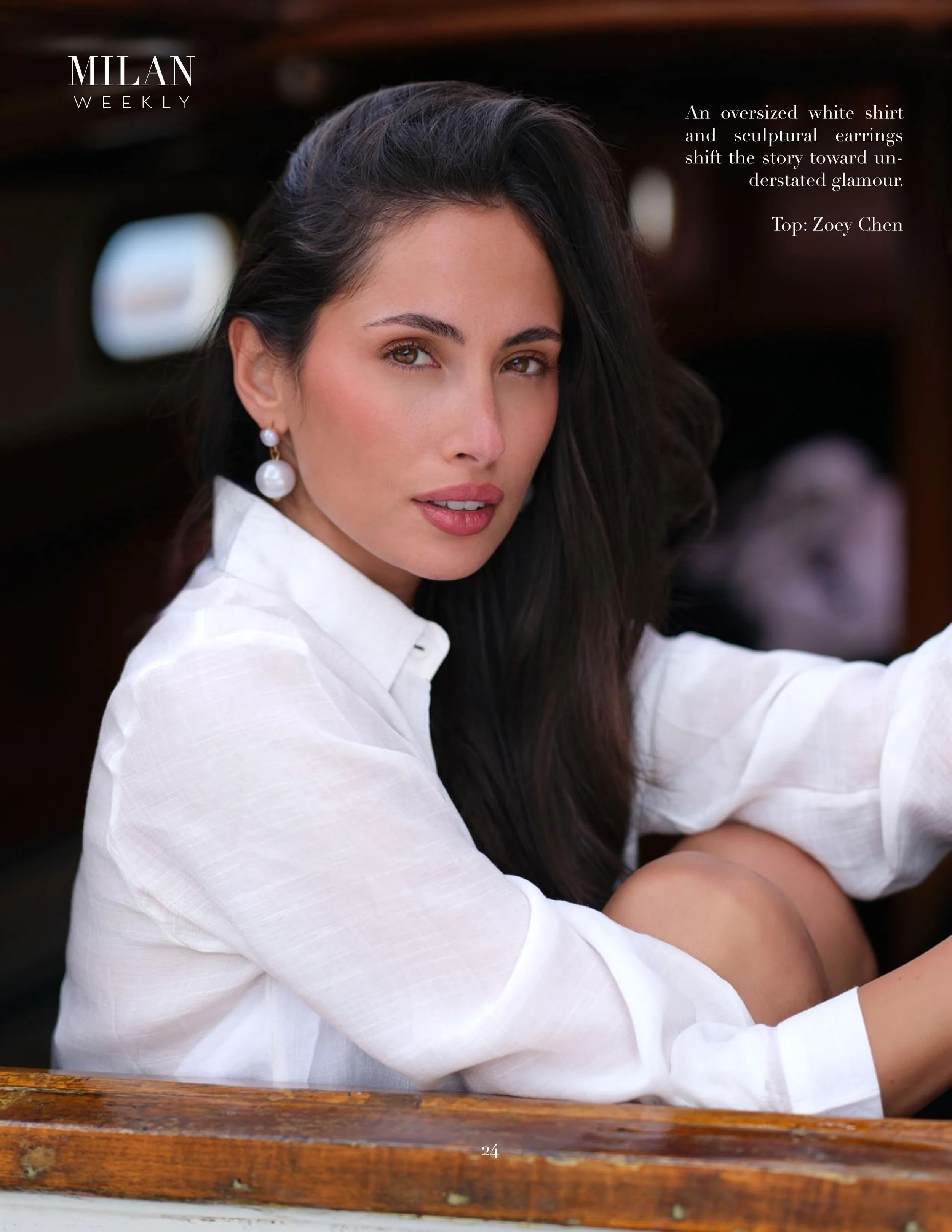 A woman with dark hair, wearing a white shirt and pearl earrings, sitting outdoors at a wooden table.