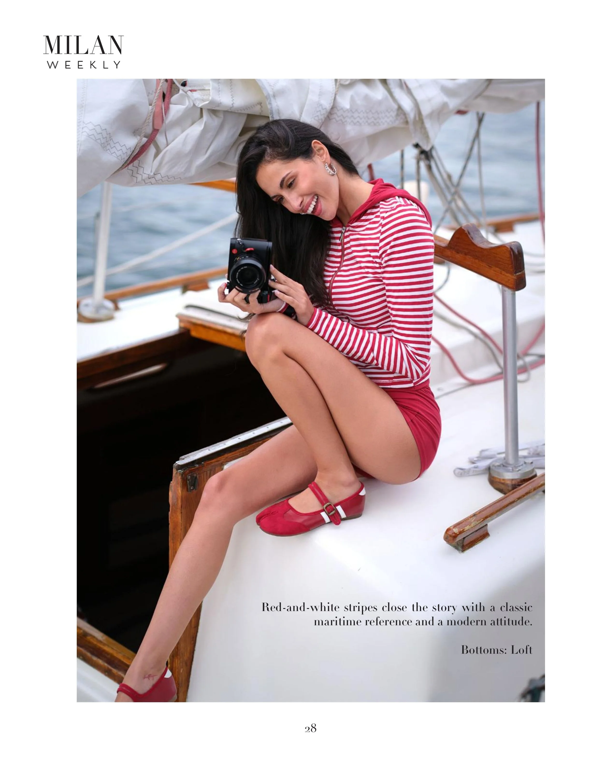 A woman with long dark hair poses on a sailboat, wearing red and white striped clothing and red shoes, smiling while looking at a camera in her hand.