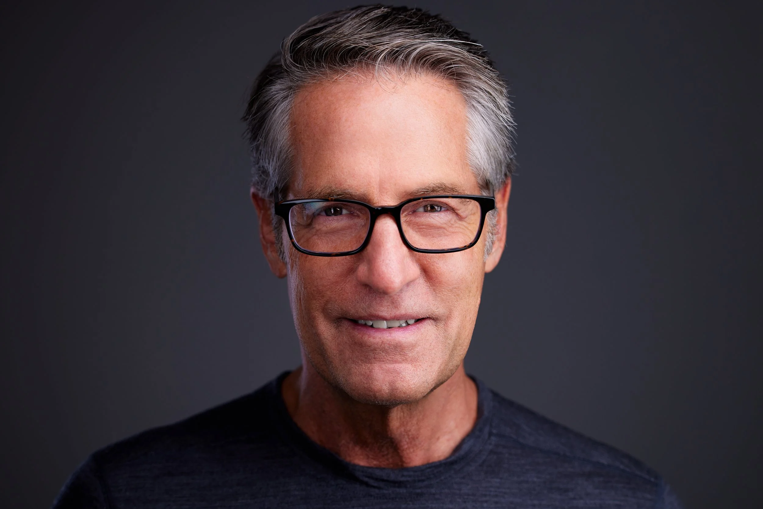 A middle-aged man with salt-and-pepper hair and glasses, smiling at the camera against a dark background.