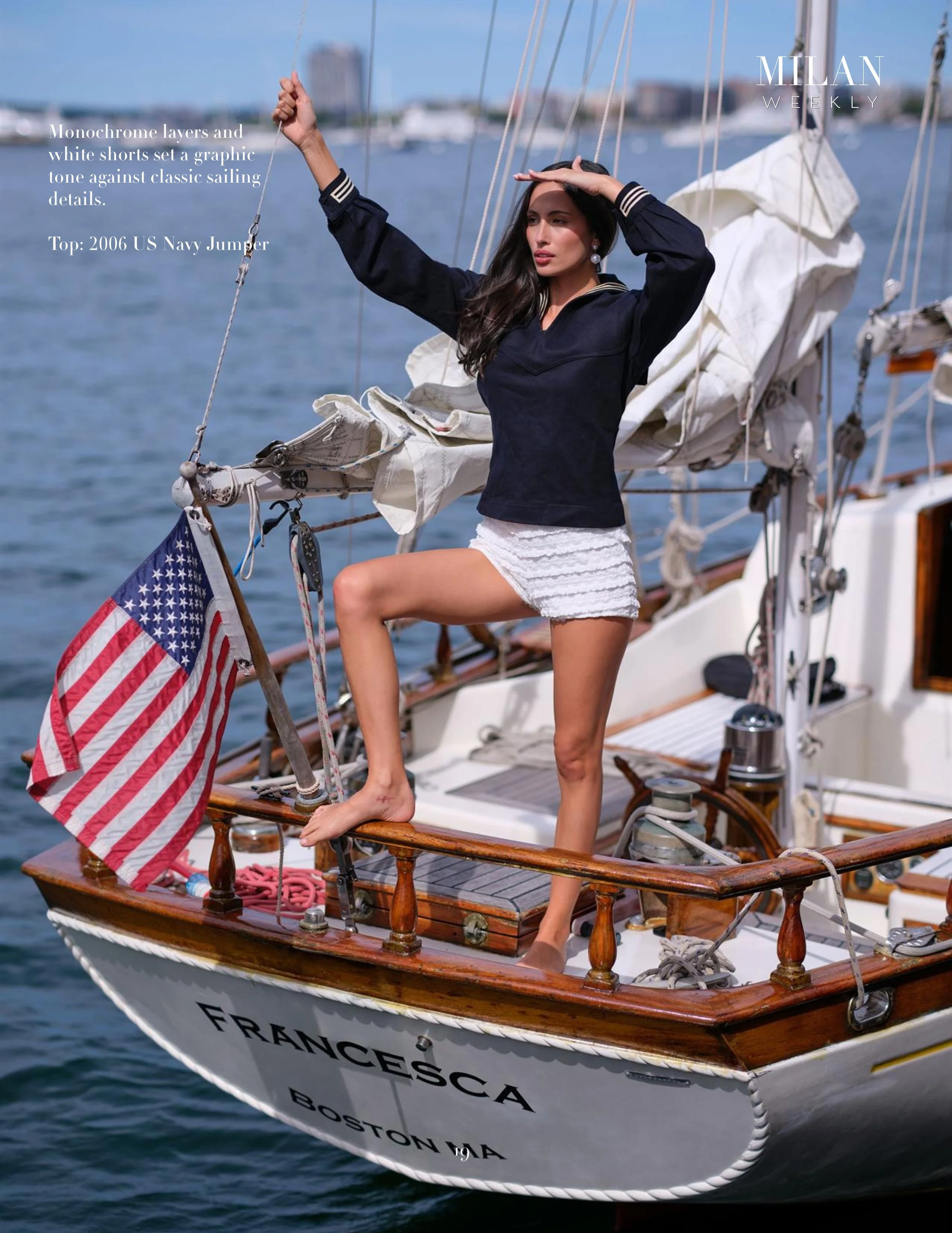 A woman standing on the deck of a boat, dressed in a navy sailor top and white shorts, saluting with her right hand while holding onto a line with her left hand. The boat has an American flag and is decorated with nautical elements. The water and cit