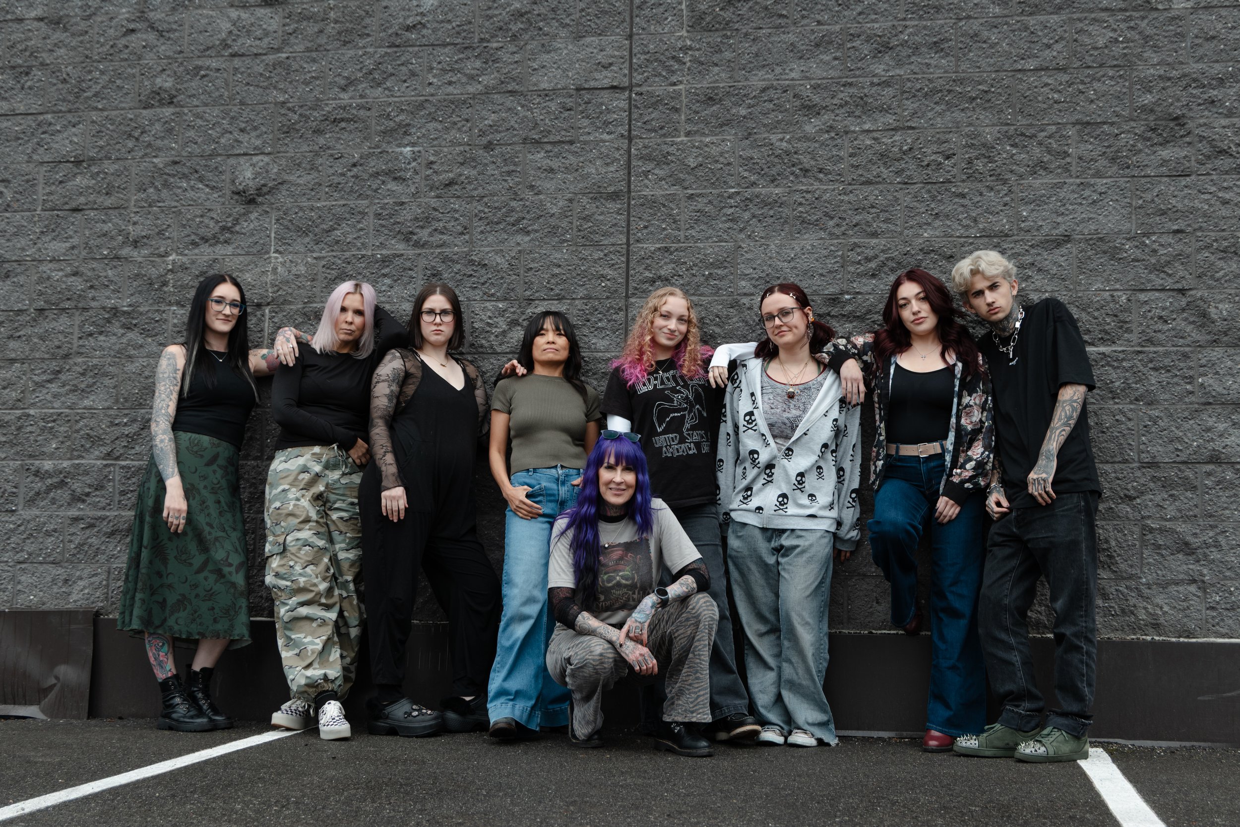A group of ten diverse young women posing closely together outdoors against a gray brick wall. Custom balayage and dimensional hair color at Misfits Salon in Anchorage, Alaska, featuring highlights, lived in blondes, seamless color blends.