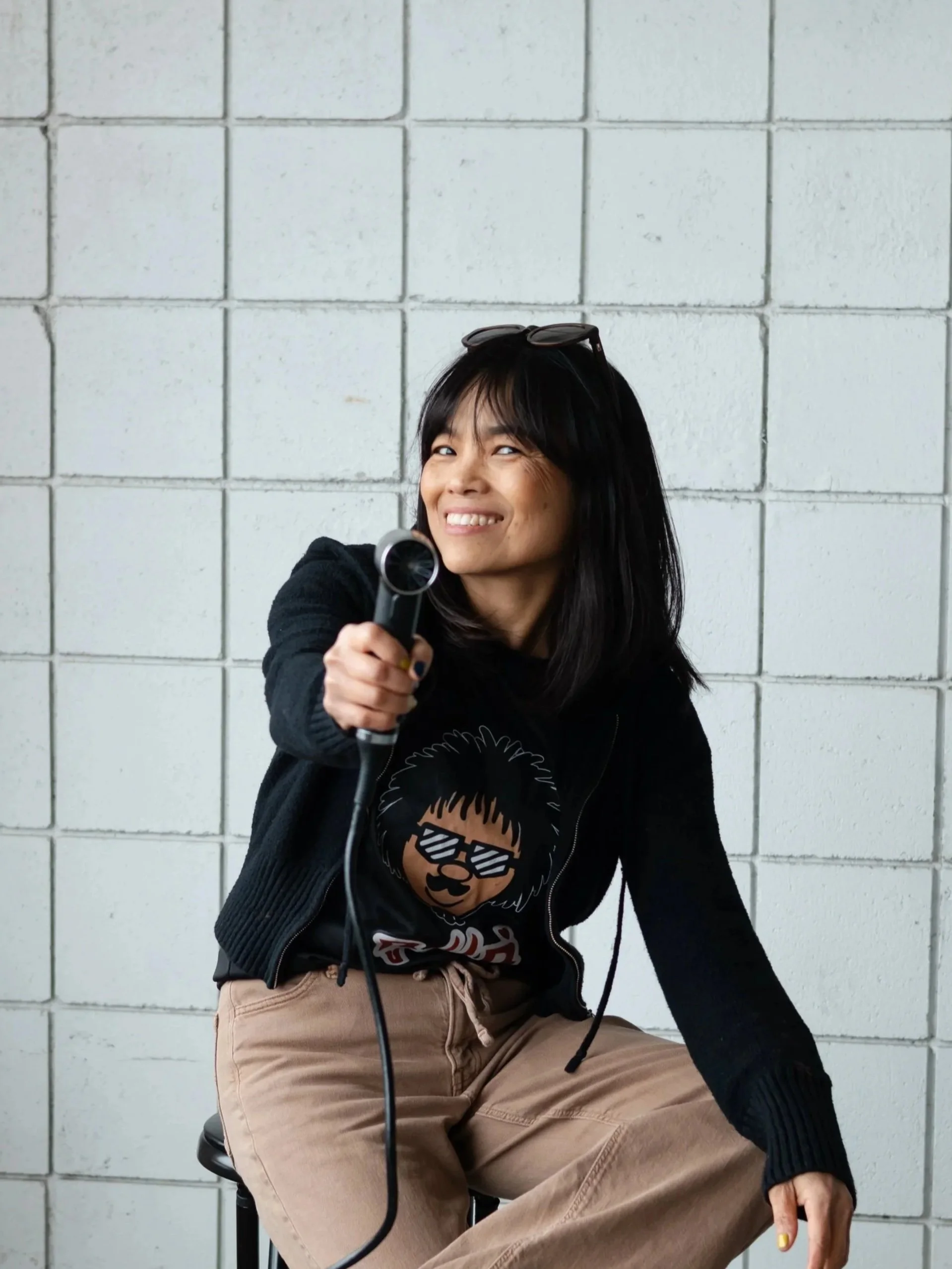 Anchorage Stylist Nee holding  blow-dryer while sitting on a stool. Nee works at Anchorage luxe Salon called Misfits Salon. She specializes in balayage, hair highlighting, blonding and ladies haircuts.